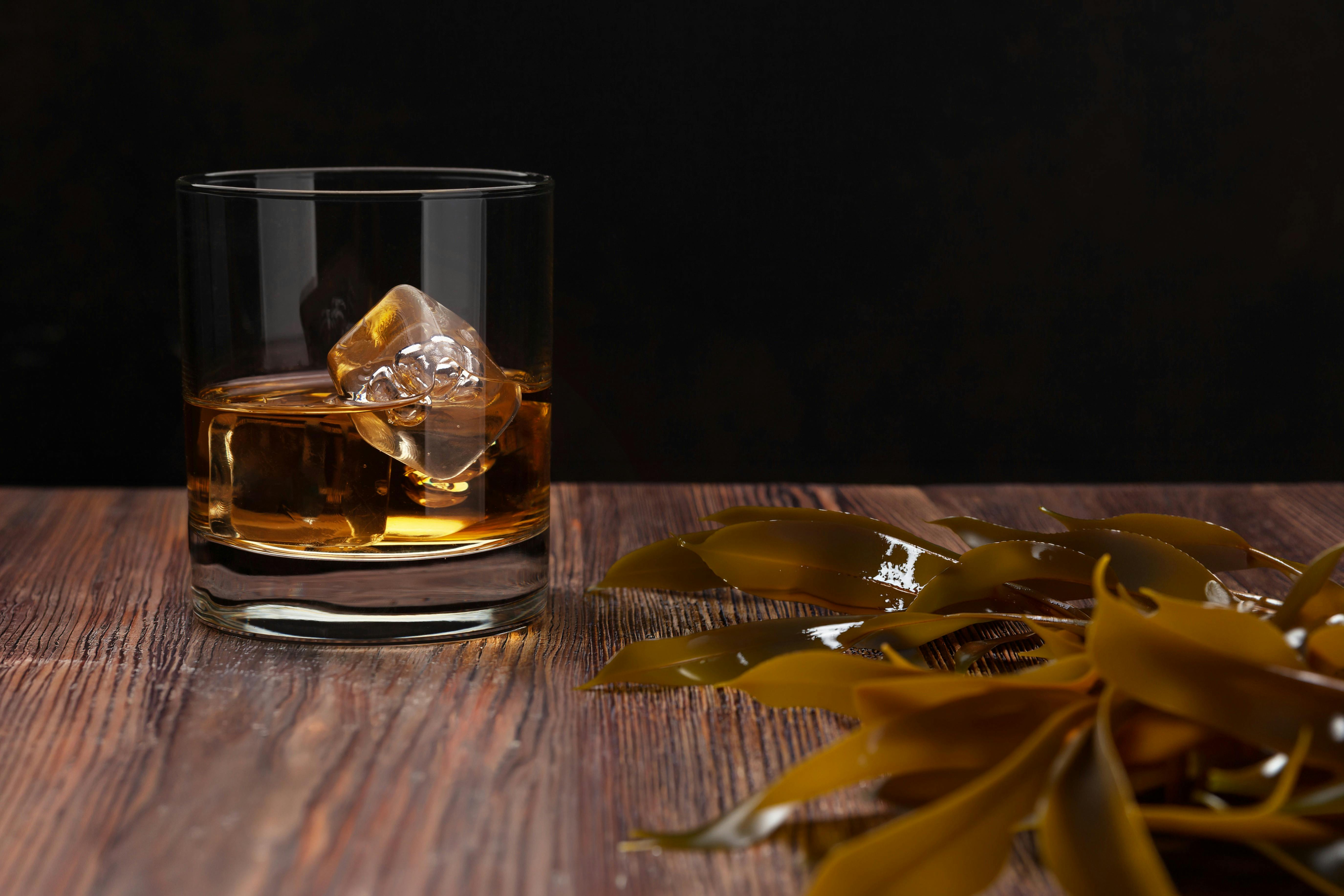 A glass of whisky sits on a wooden table alongside brown kelp, reflecting the colour of the drink