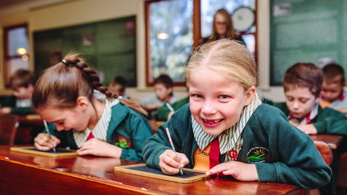 Little primary school girl sitting at a desk with a cheeky smile as she write on a slate