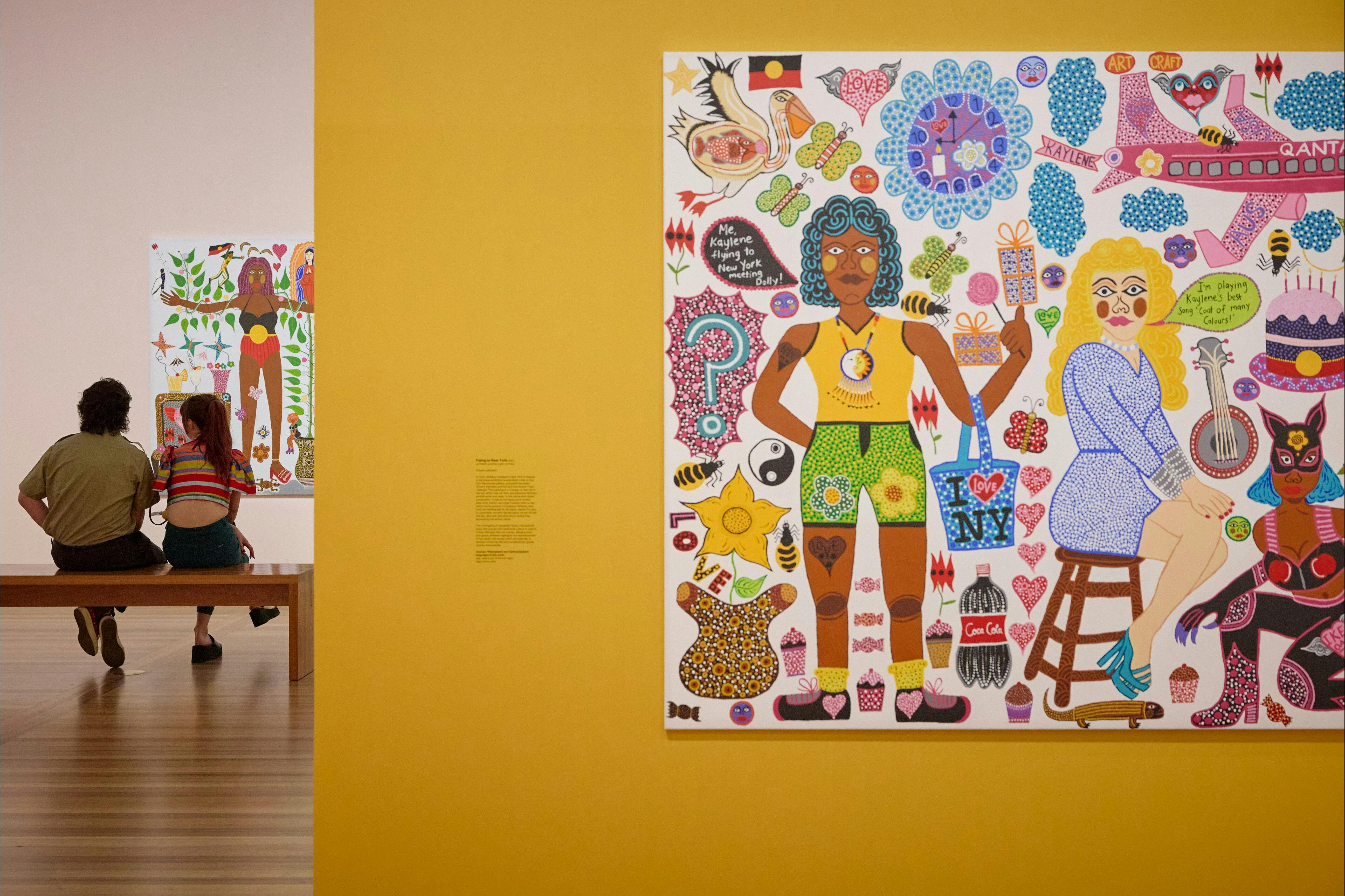 Two young people view a portrait, and in the foreground, a colourful portrait hangs on a yellow wall