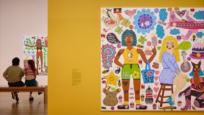 Two young people view a portrait, and in the foreground, a colourful portrait hangs on a yellow wall