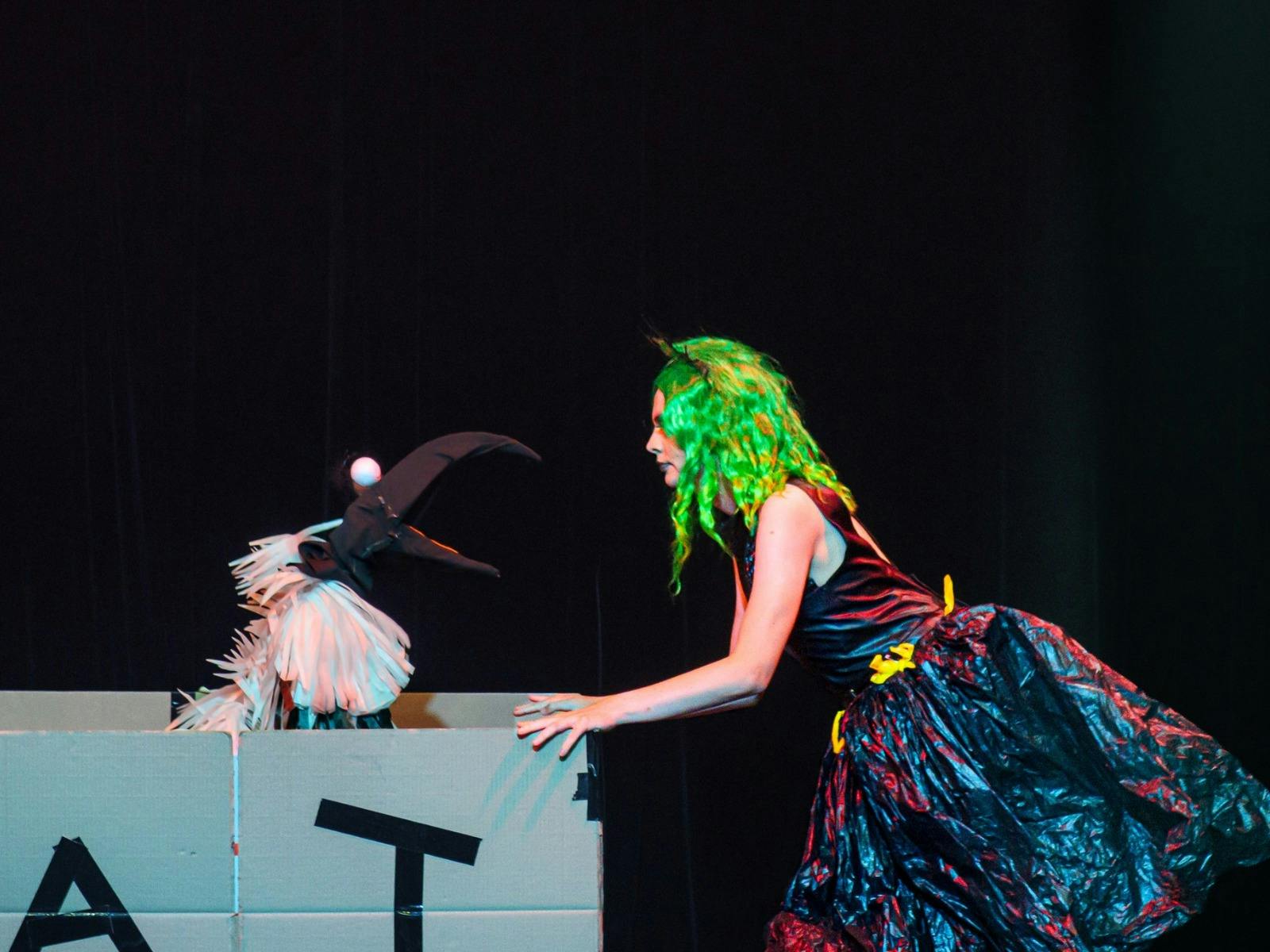 A drag queen green in garbage bags and a green wigs looks at a ibis puppet