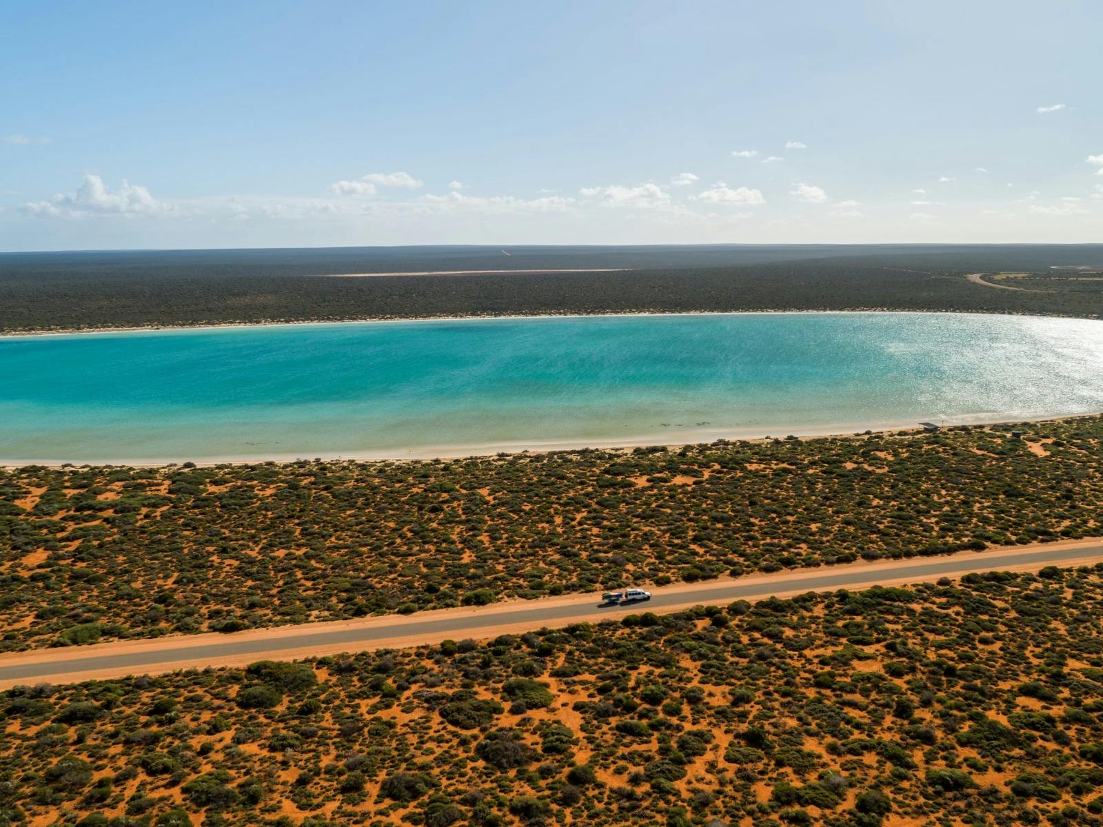 A holiday caravan driving through a red desert landscape, passing Lake Thedham in Shark Bay