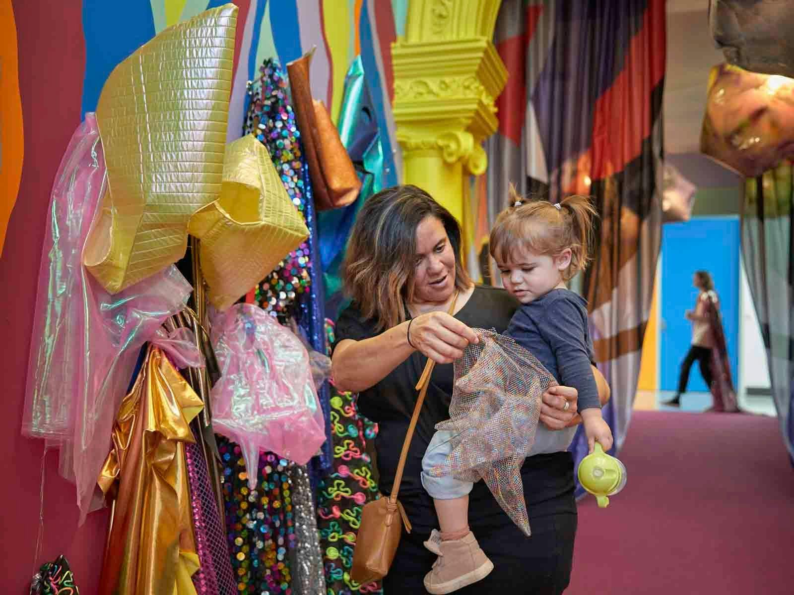 Mother and toddler look at glittering costumes in a brightly painted gallery