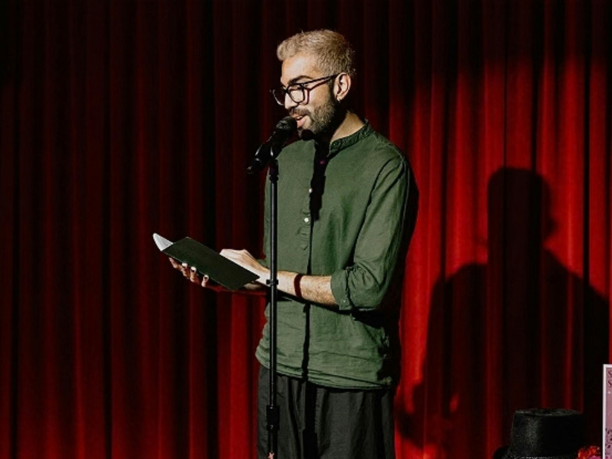 A poet reads into a microphone against a red velvet curtain, wearing a green shirt & black pants.