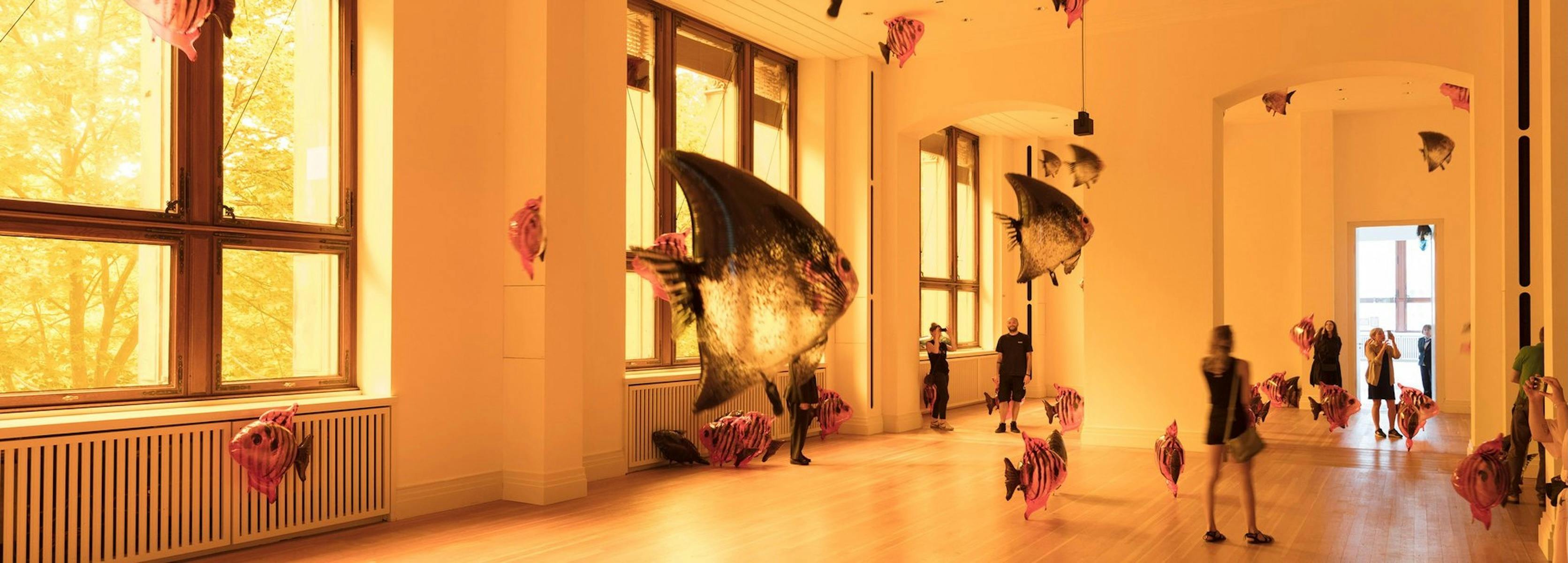 A sunlit museum gallery glows amber as visitors walk among oversized suspended fish sculptures