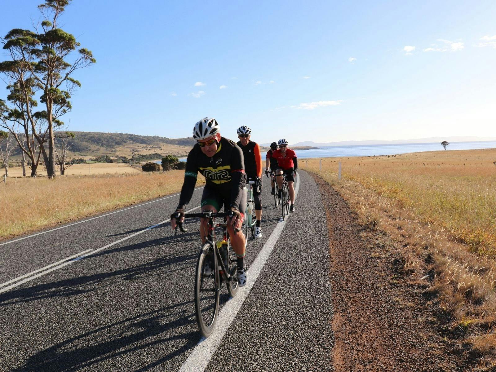 Cyclists riding on road with clear blue sky and ocean behind them.
