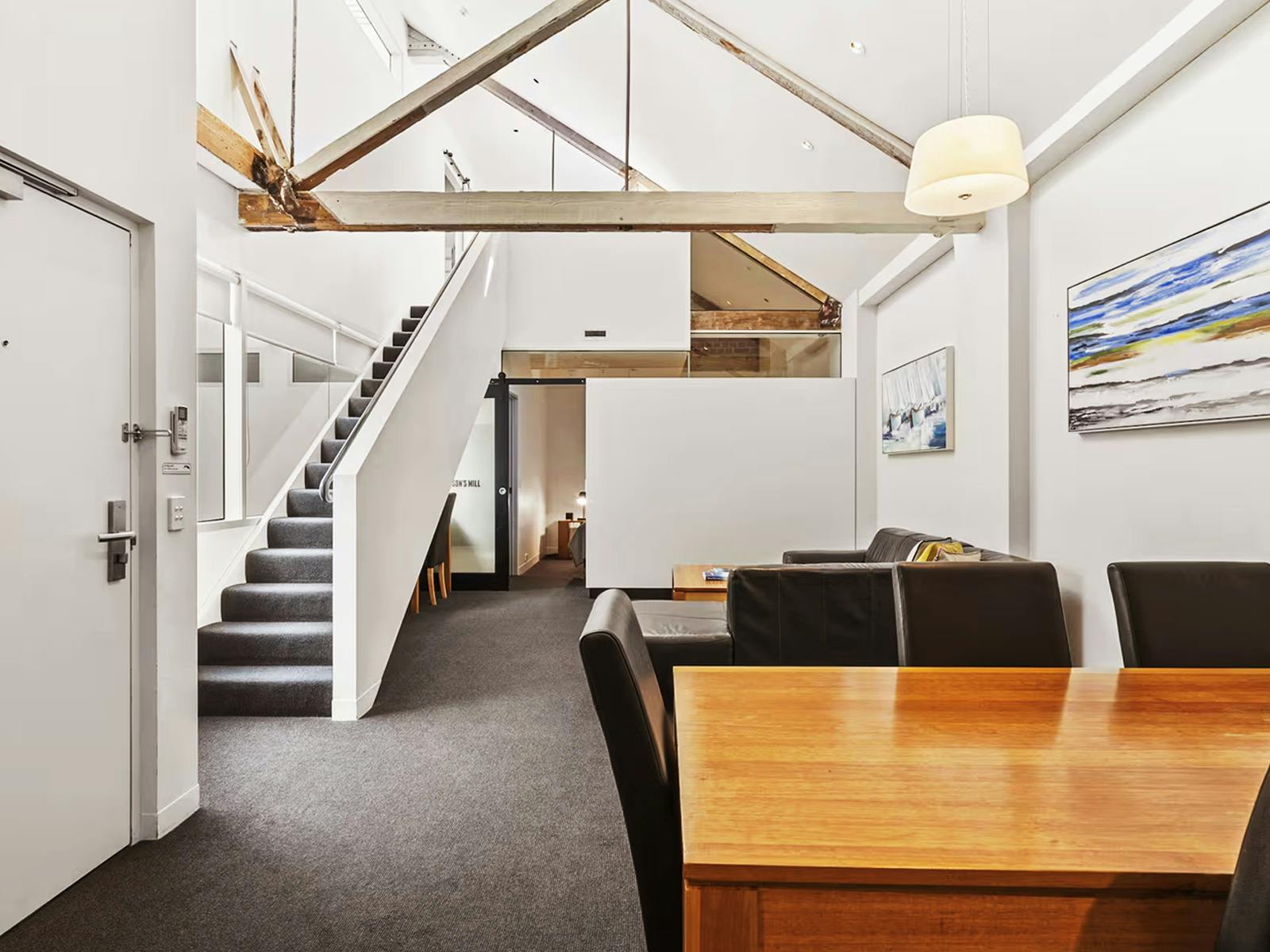 Split-level apartment hallway with timber staircase, dining table, and exposed roof beams.