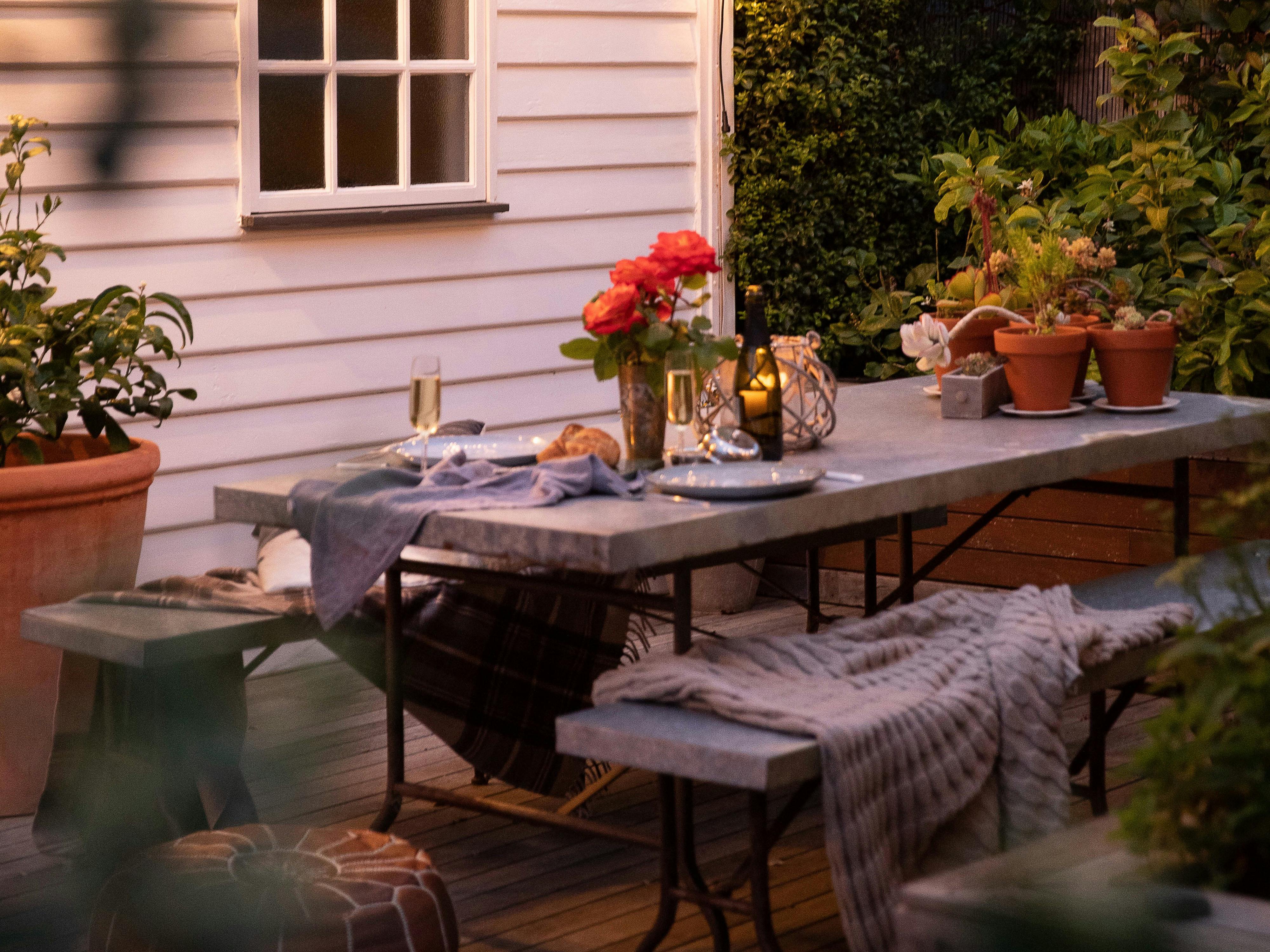 decked courtyard with an outdoor dining table, surrounded by greenery, lit with festoon lighting.