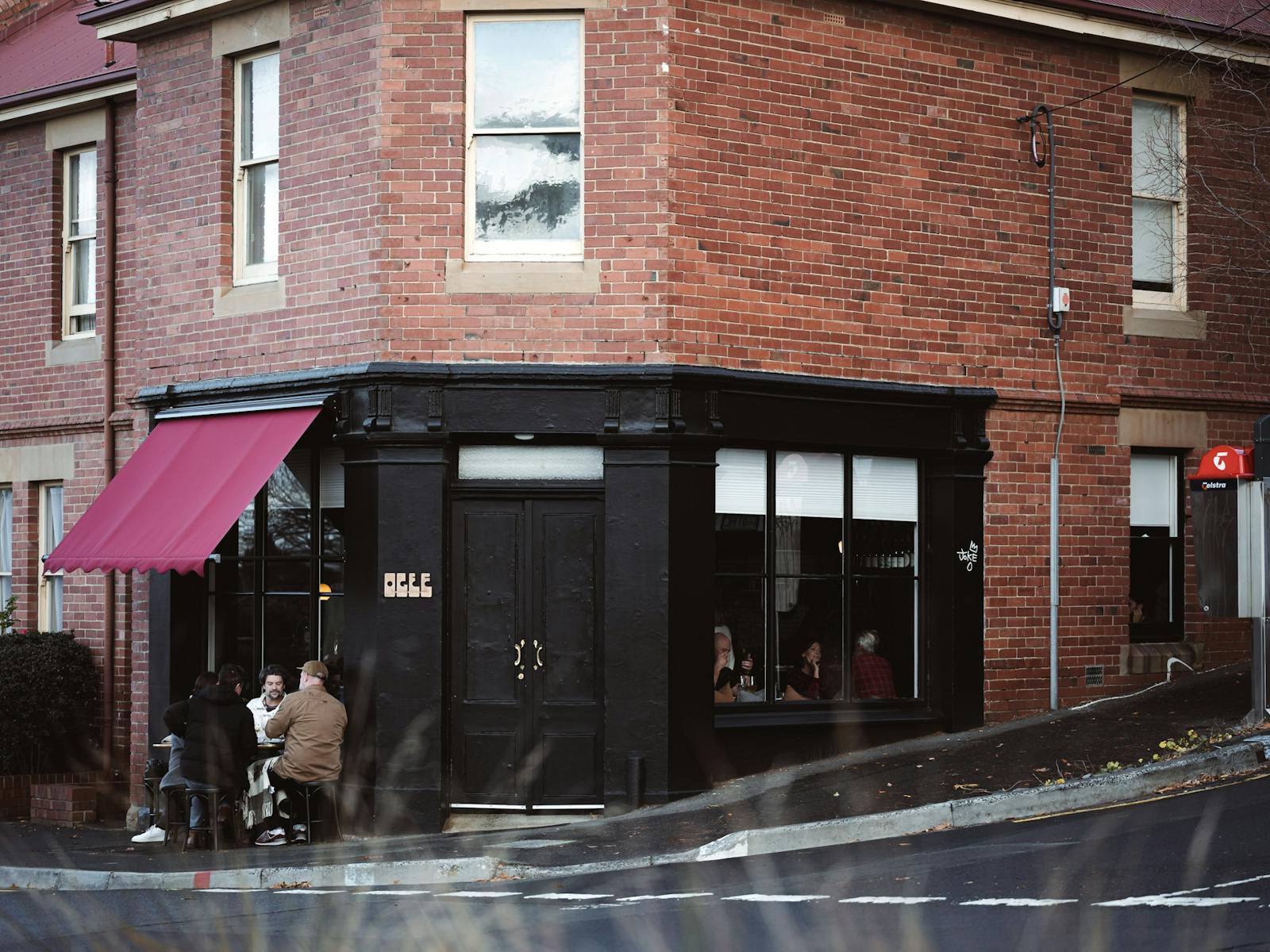 Photo of the exterior of Ogee. Black corner shop with red awning. People enjoying a drink out front.