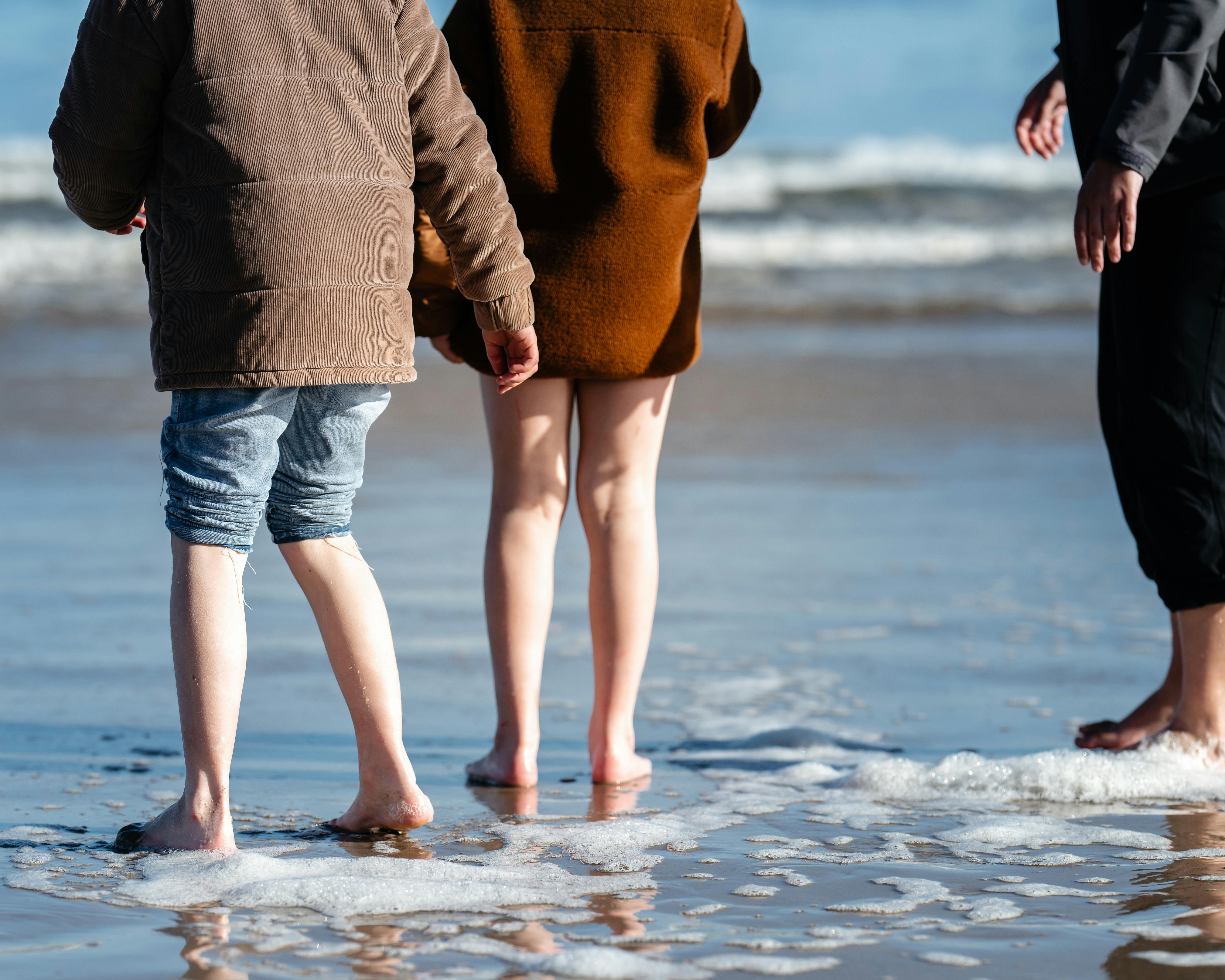 Three people stand barefoot on the wet sand at the beach, with waves gently lapping around their fee