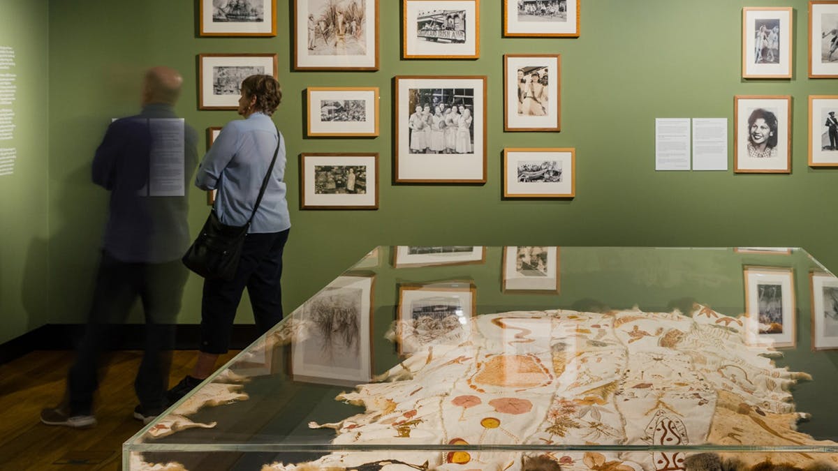 Two people standing in a green-walled gallery space looking at pictures hanging. Two people standing in a green-walled gallery space looking at pictures hanging.