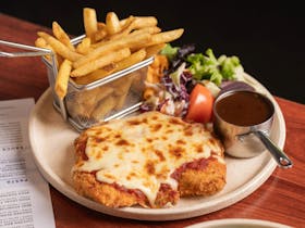 A plate with a chicken parmigiana, a basket of hot chips, some salad and a vessel of gravy.