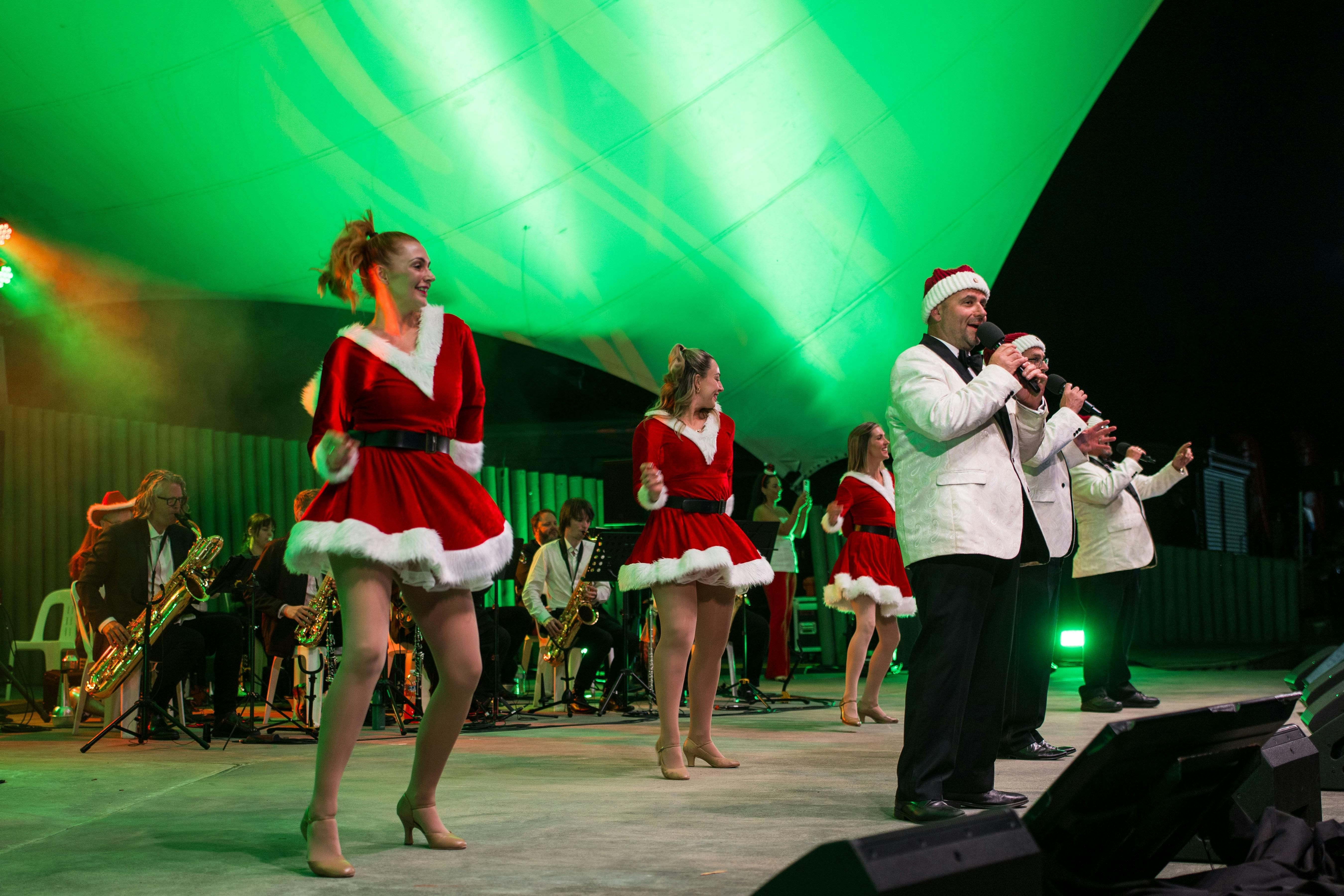 Peformer John X  in a santa hat singing with dancing girls in santa suits