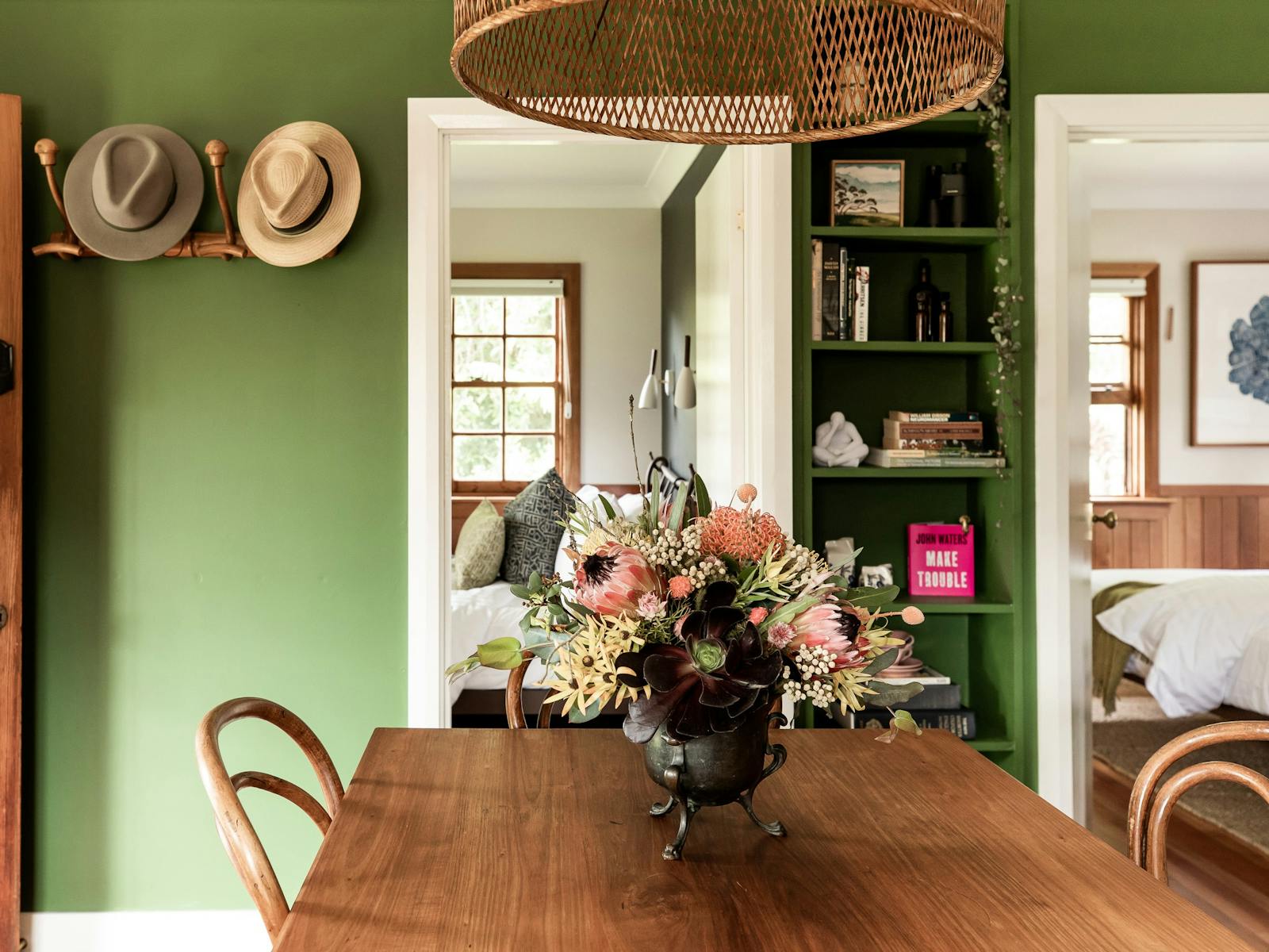 Wooden dining table with a vase of native flowers sits in front of a green wall with book alcove