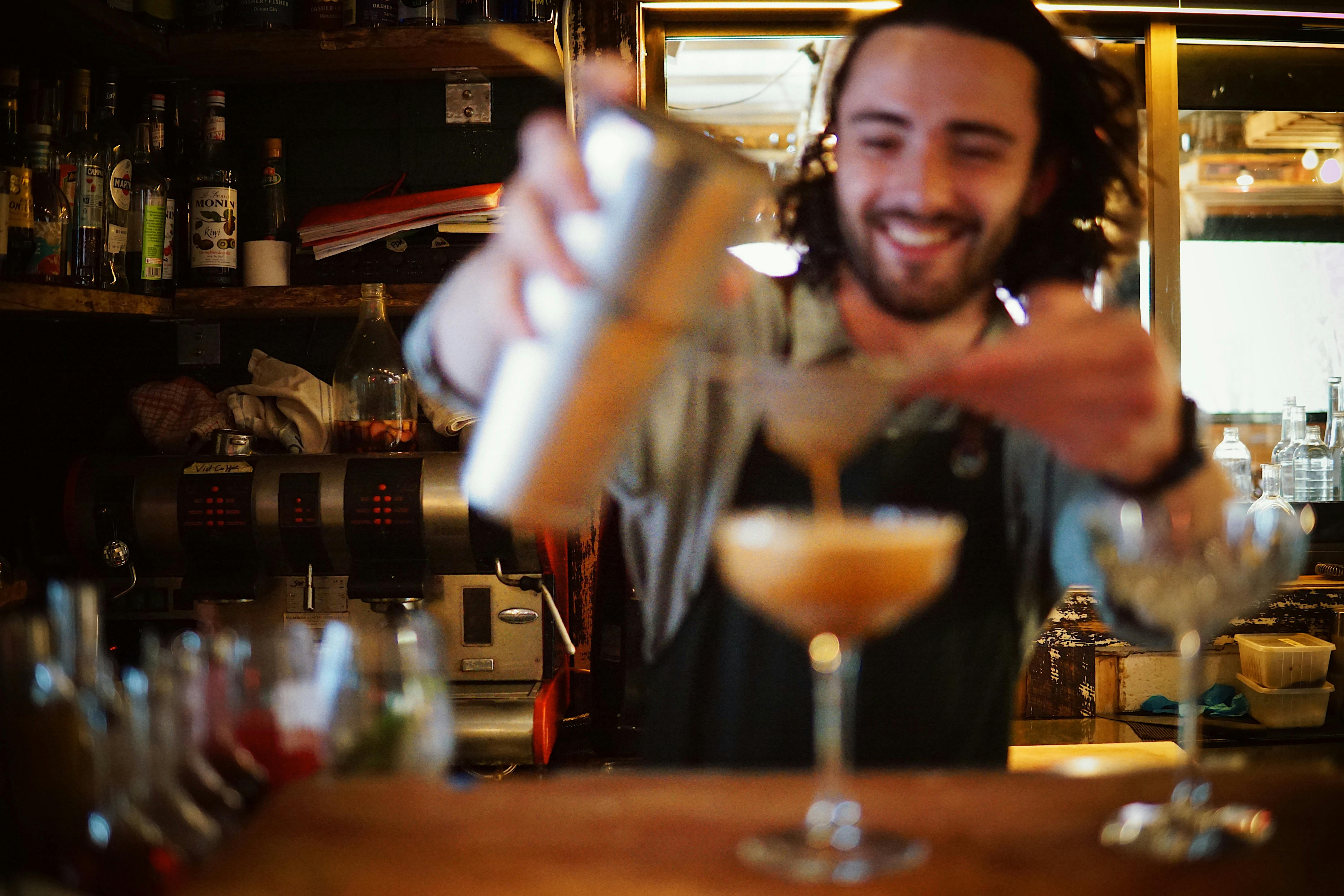 Bartender is smiling in focus, while out-of-focus pours liquid from a cocktail shaker into a coupe