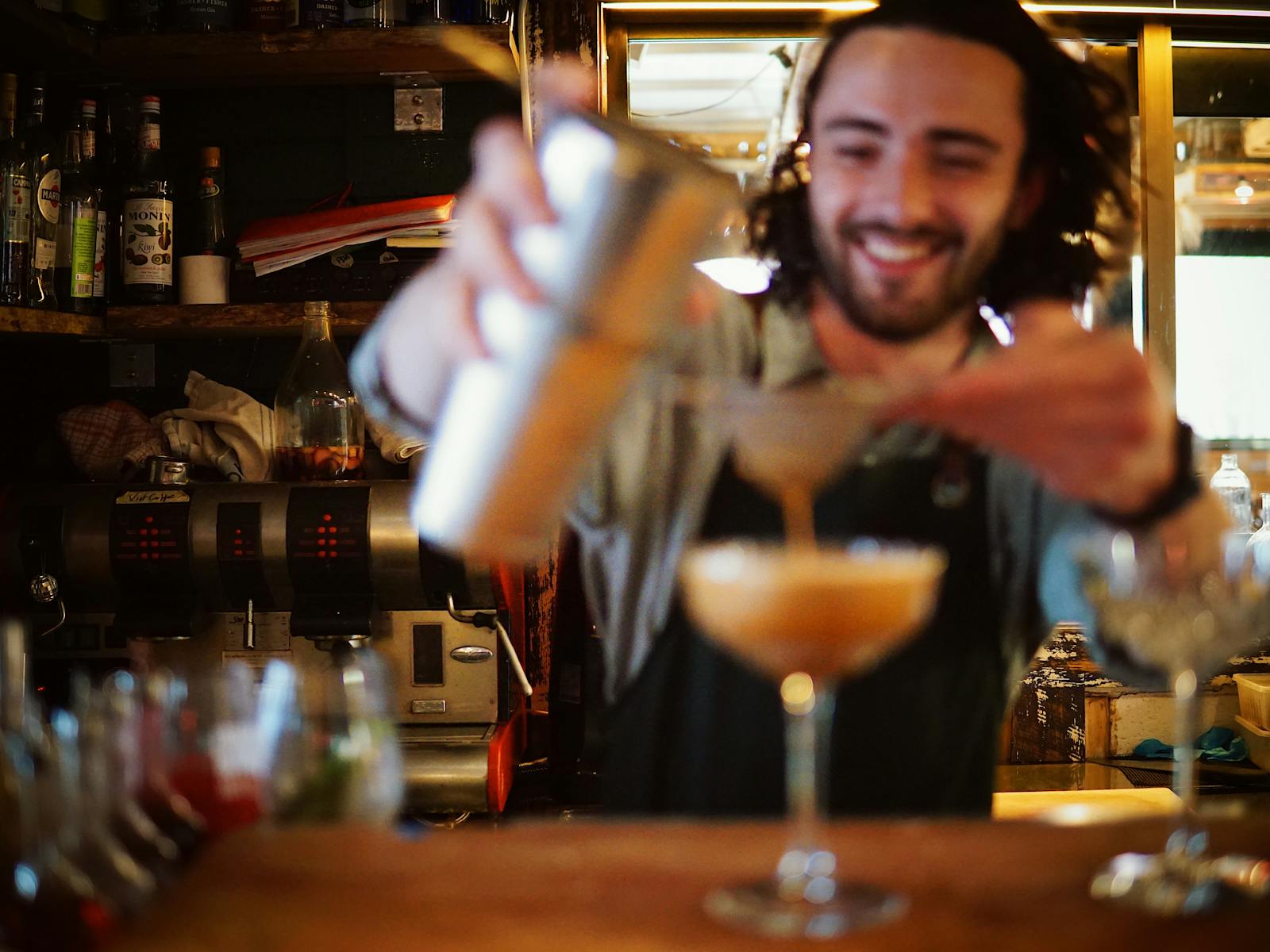 Bartender is smiling in focus, while out-of-focus pours liquid from a cocktail shaker into a coupe