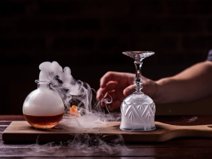 A smoking bottle on a wooden board with a hand about to turn over a smoke filled glass