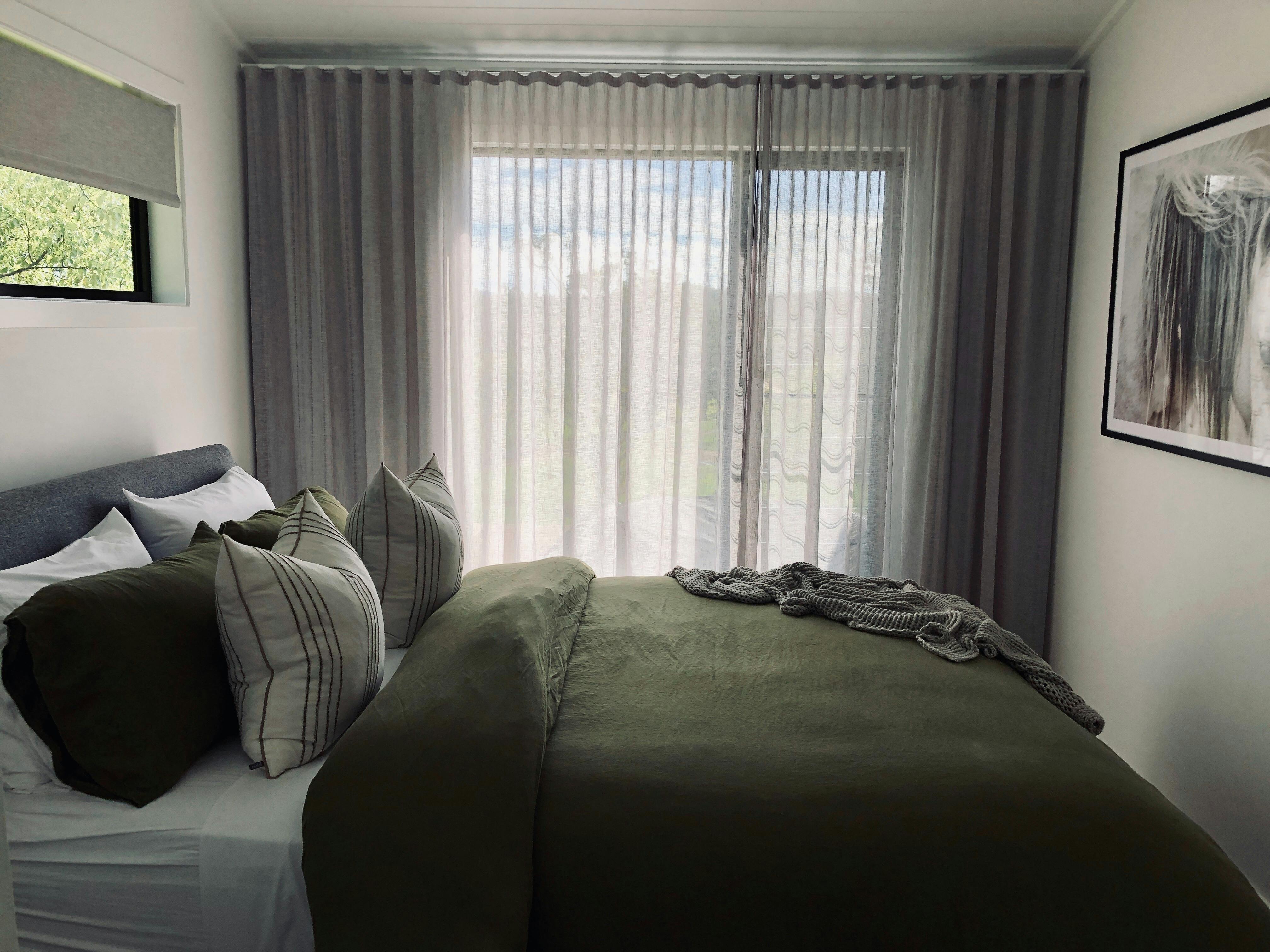 퀸 사이즈 침대에 녹색 리넨. 쌓인 쿠션과 러그. 큰 창을 통해 여과된 빛.