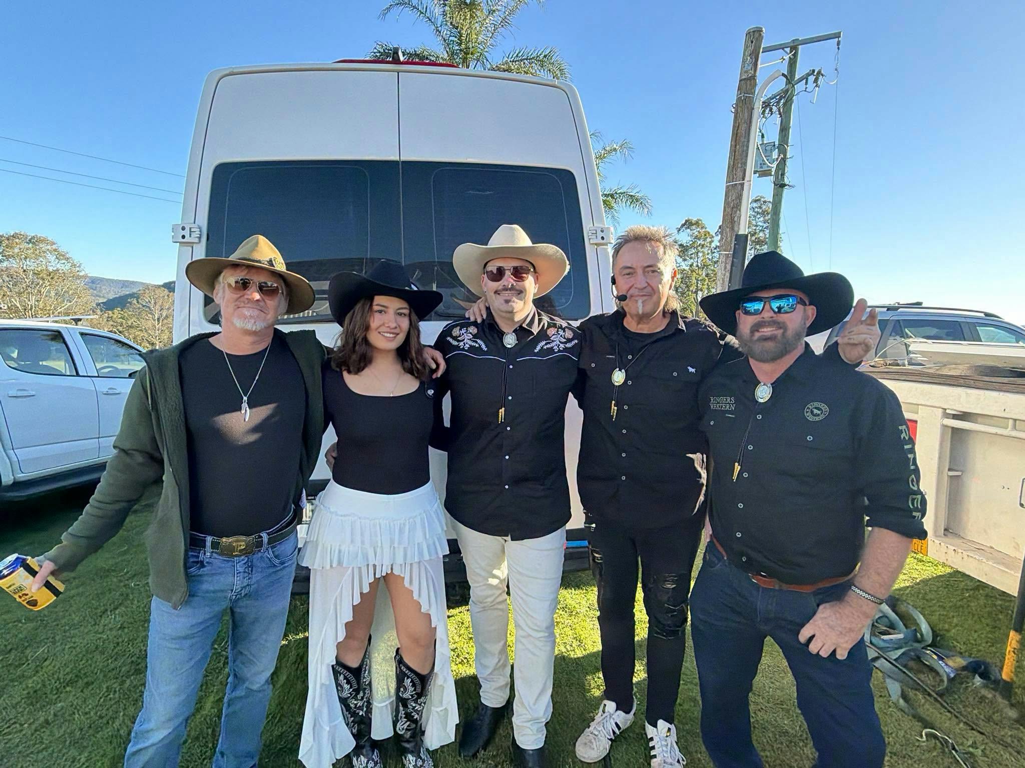 The Dirt Road Tribute Band poses together in front of their tour bus on a bright sunny day, wearing