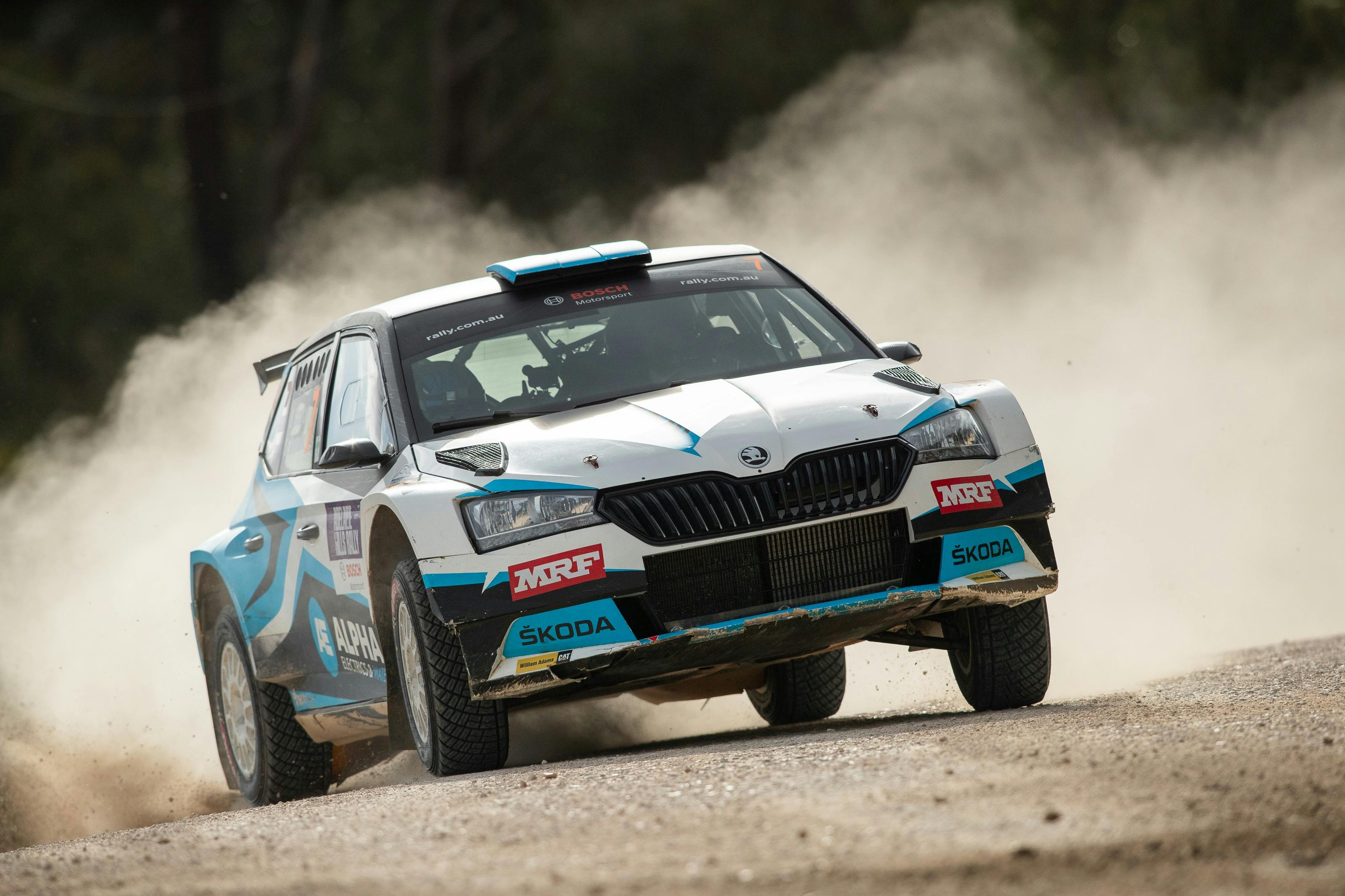White and blue rally car is driving on a gravel road with dust kicked up behind