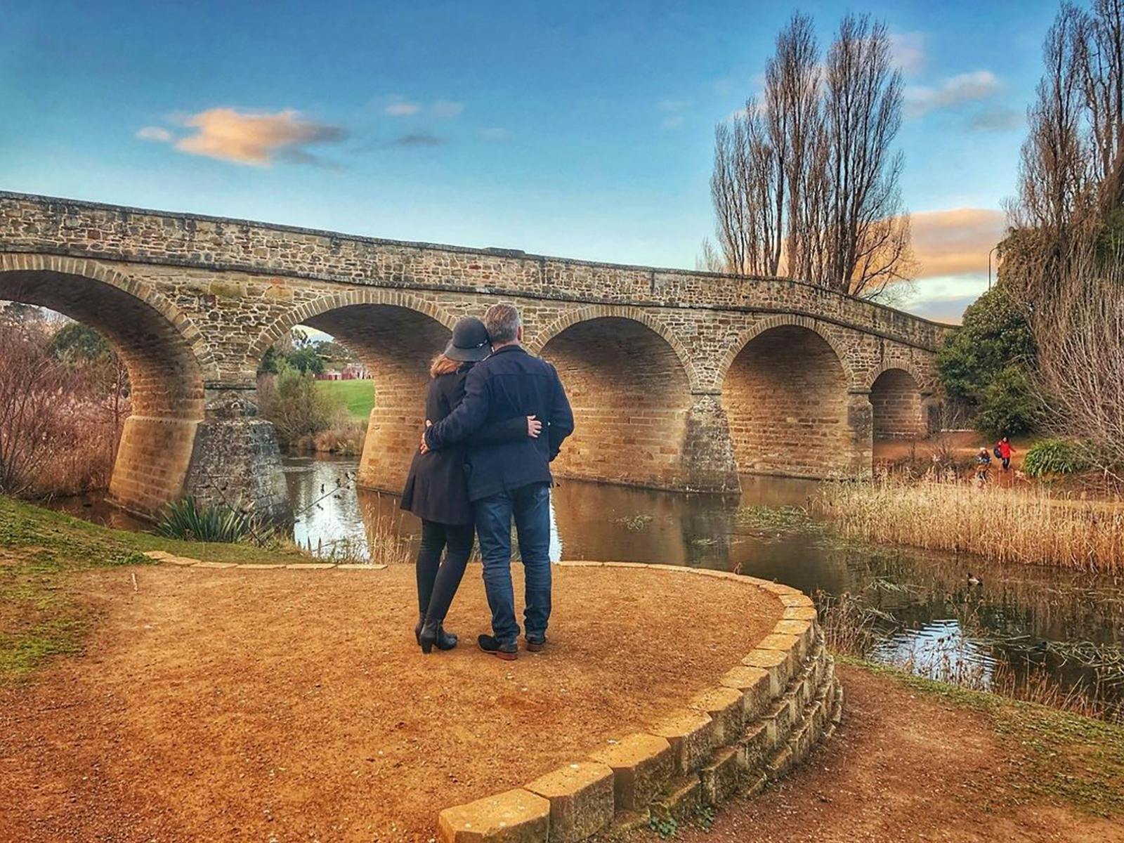 "Romantic couple embraces in front of Richmond Bridge, enveloped in serene solitude. Their tender mo