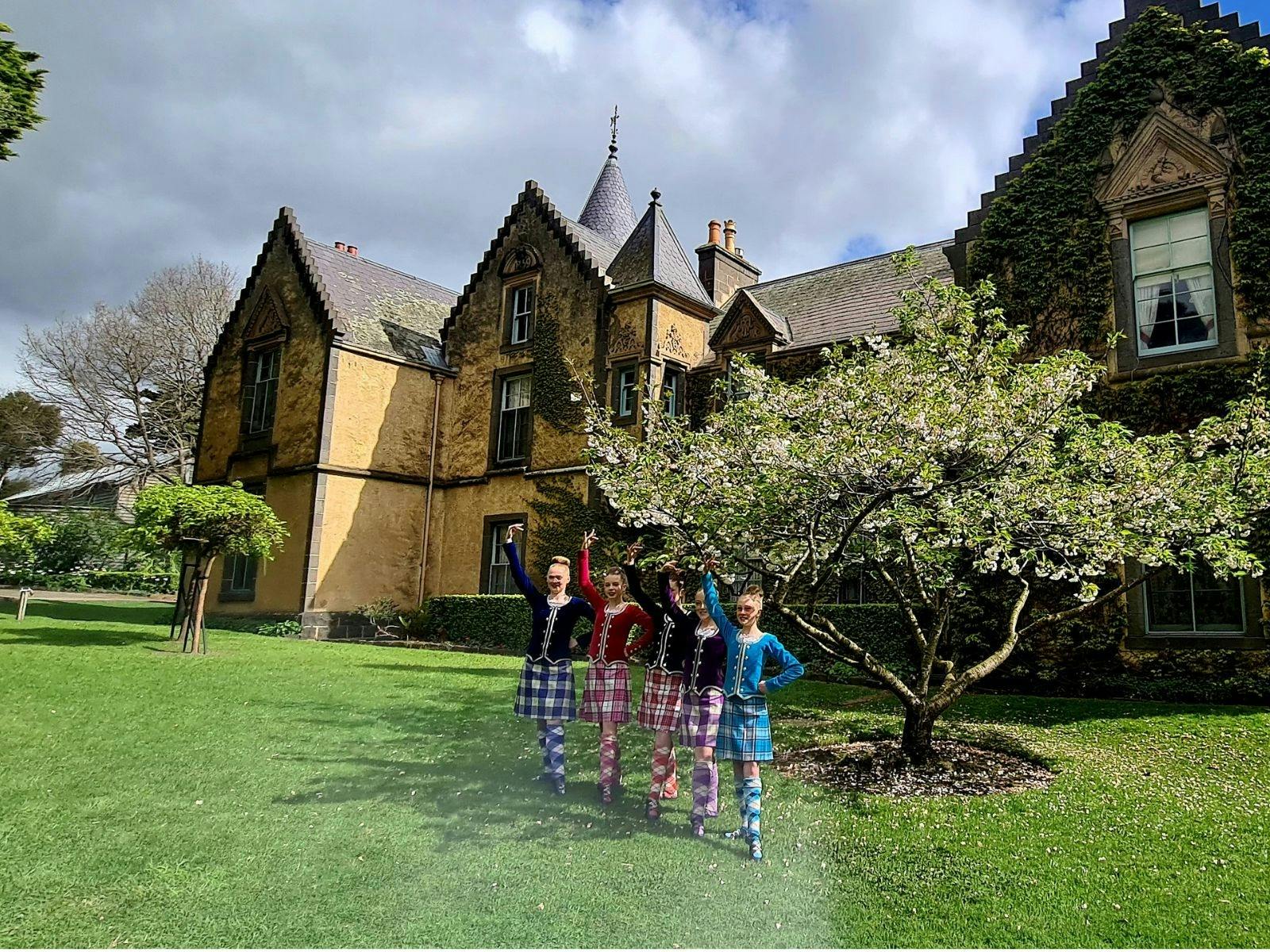 Five girls in Scottish attire stand in front of a tree, with Overnewton Castle (a large manor) behin