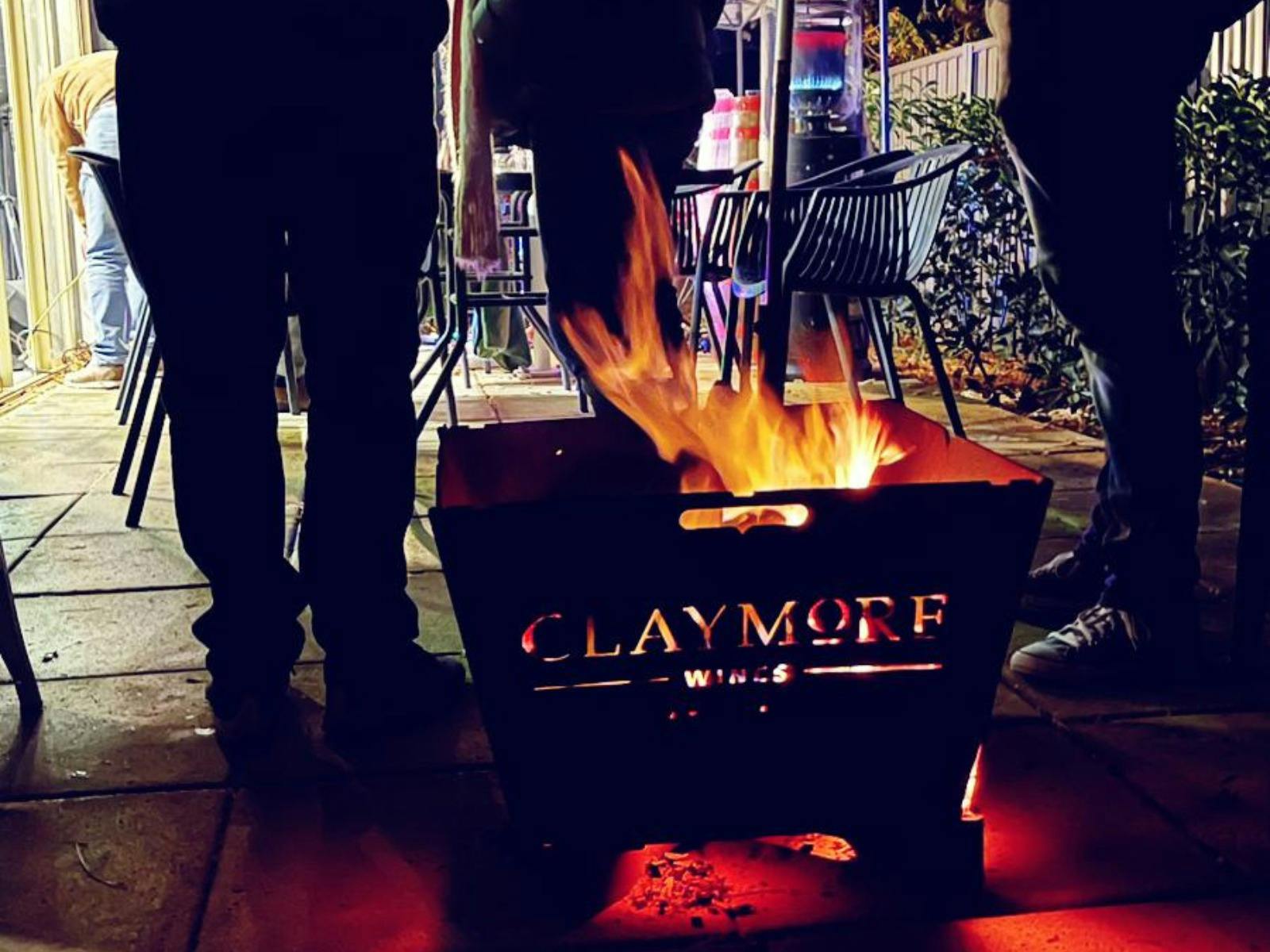 an open fire pit branded with Claymore in front of the legs of guests at an outdoor event