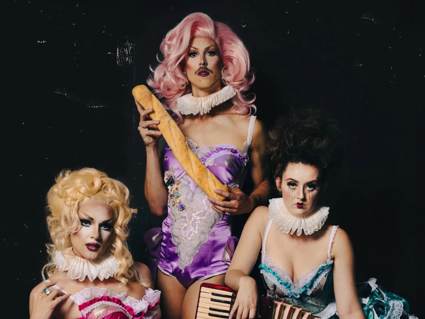 Three drag cabaret performers pose in ornate 18th-century-inspired lingerie with ruffled collars