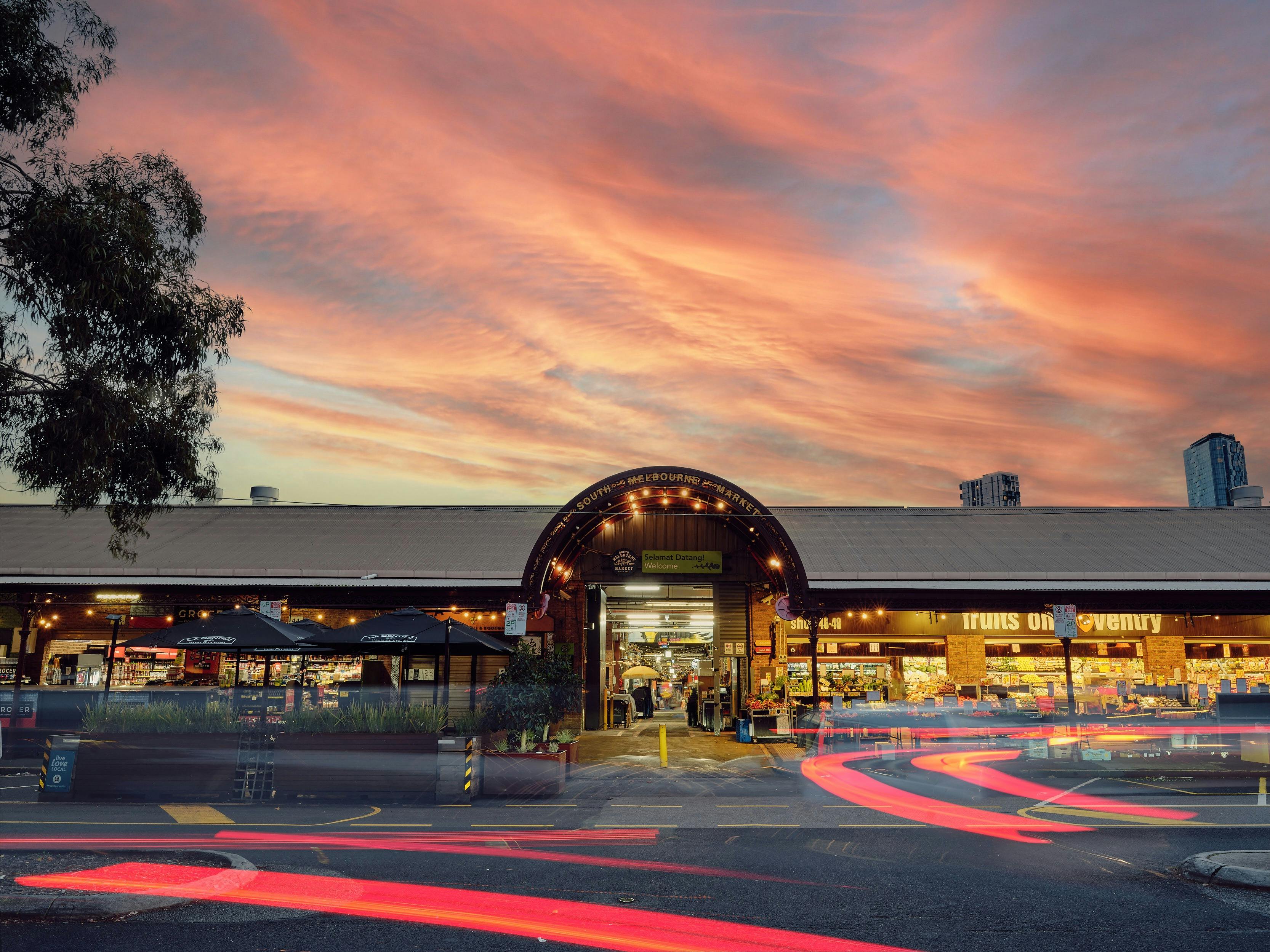 South Melbourne Market entrance at sunset, with illuminated stalls and car light trails.