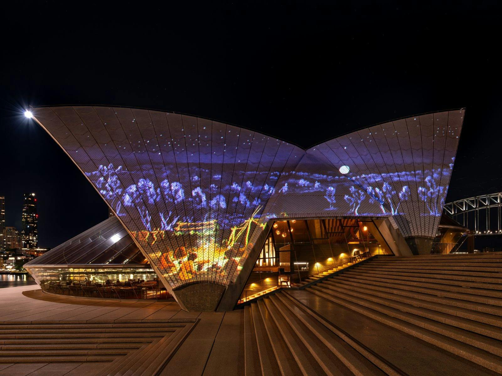 The Opera House western sails lit up with people sitting around a campfire  with a dark night sky