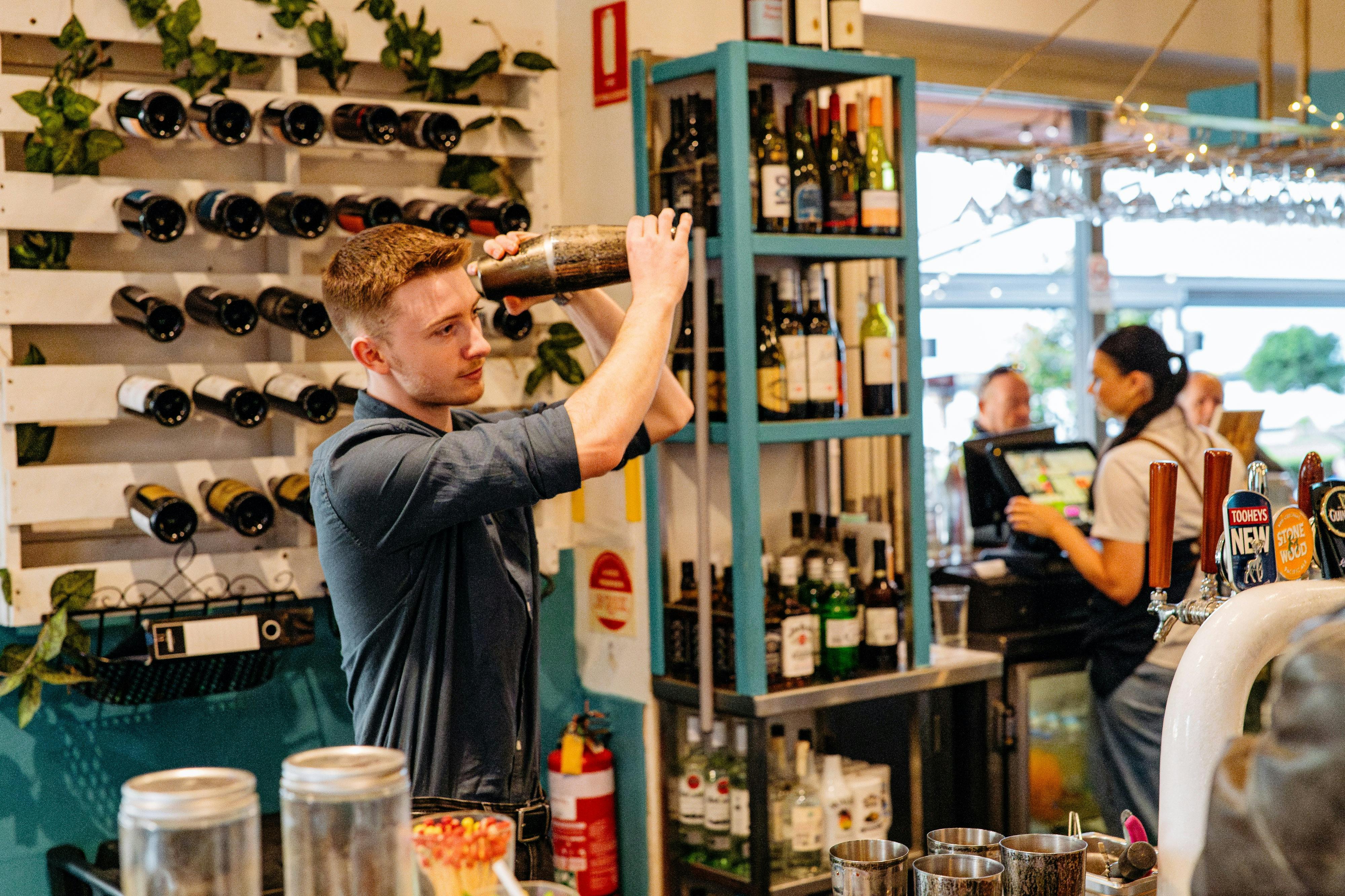 A male bartender vigorously shaking a cocktail shaker behind a well-stocked bar.