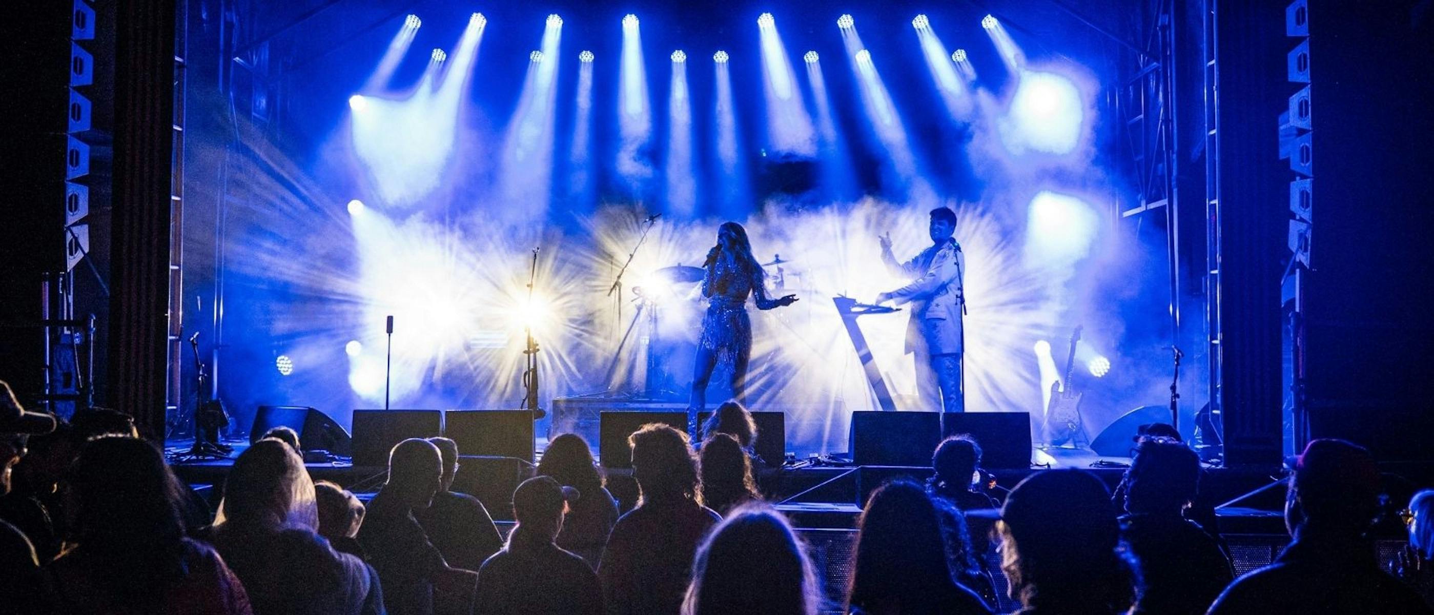 Two performers on stage in front of an audience, with bright lights behind them