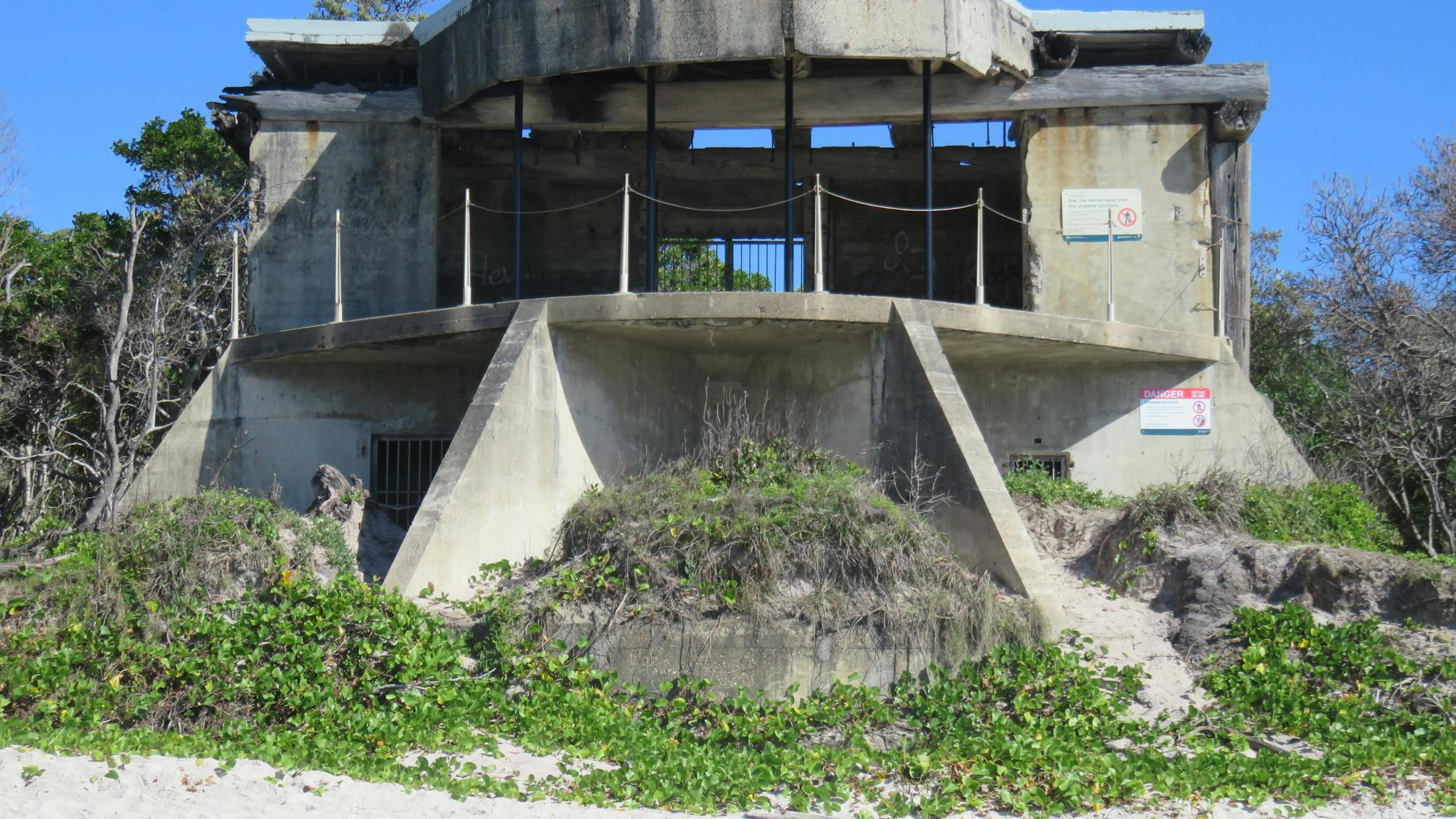 Concrete gun emplacement sits on sandy beach backed by coastal plants.
