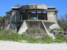 Concrete gun emplacement sits on sandy beach backed by coastal plants.