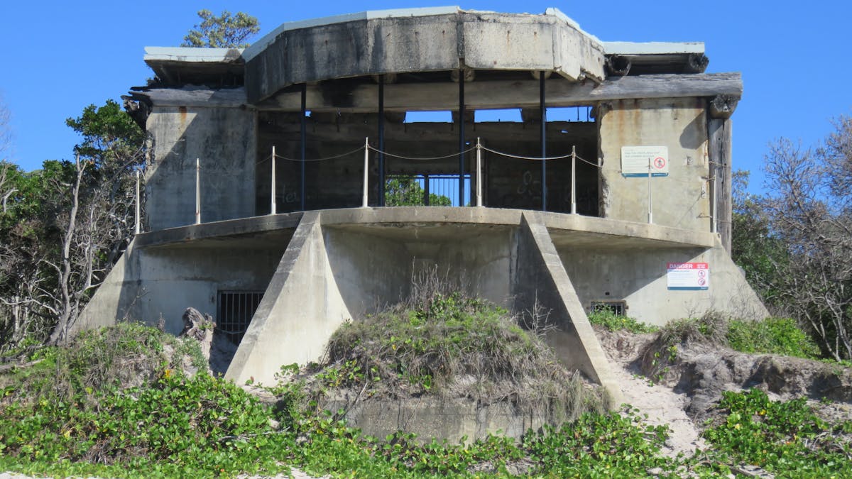 Concrete gun emplacement sits on sandy beach backed by coastal plants.
