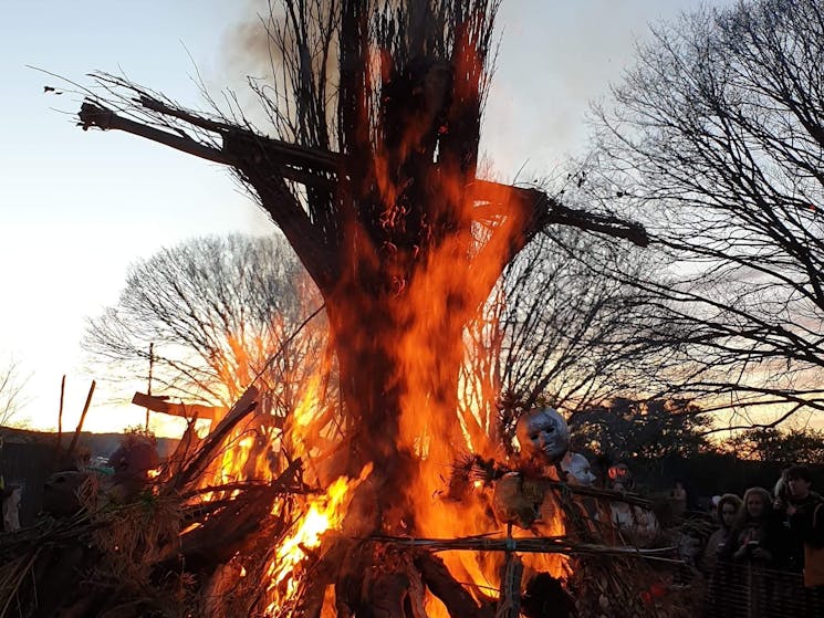 Flames are licking up the tower of the wicker man, with a flaming bonfire below, bare trees behind