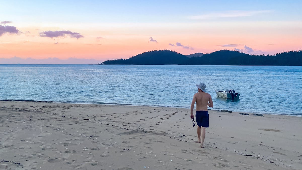 A man walking on a beach towards small boat in water and sunset over island hills in the distance