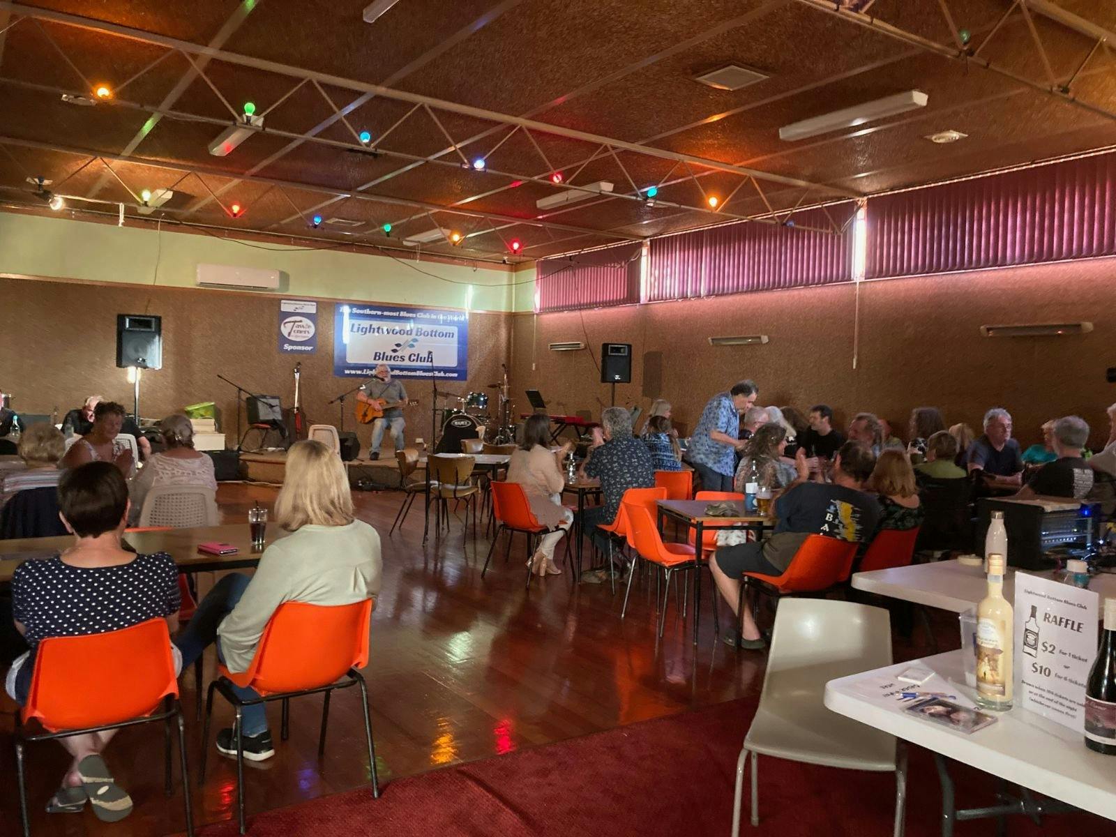 A room filled with people with a guitarist on stage in the background