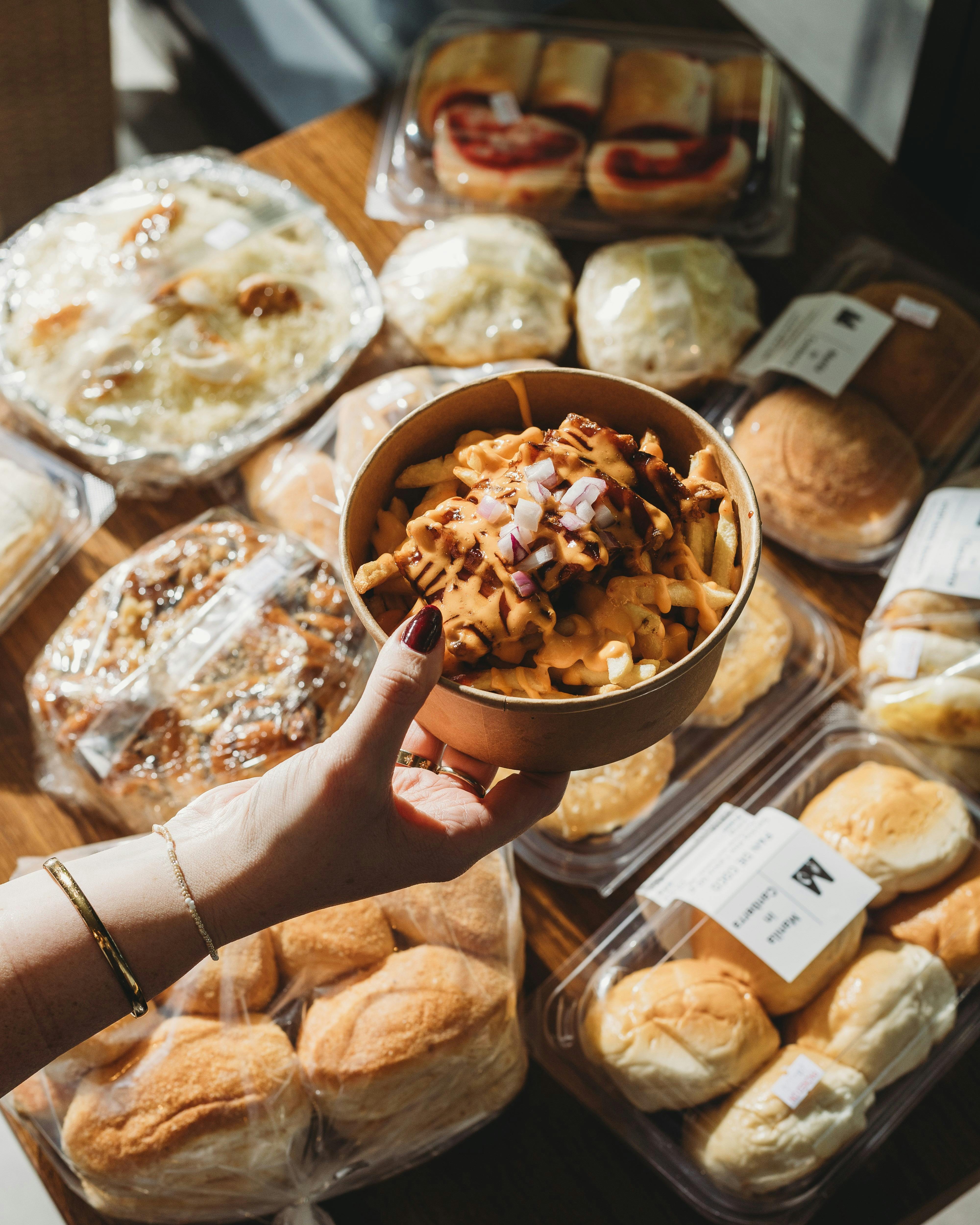 A hand holding a bowl of lechon loaded fries topped with cheese and onions, surrounded by assorted F