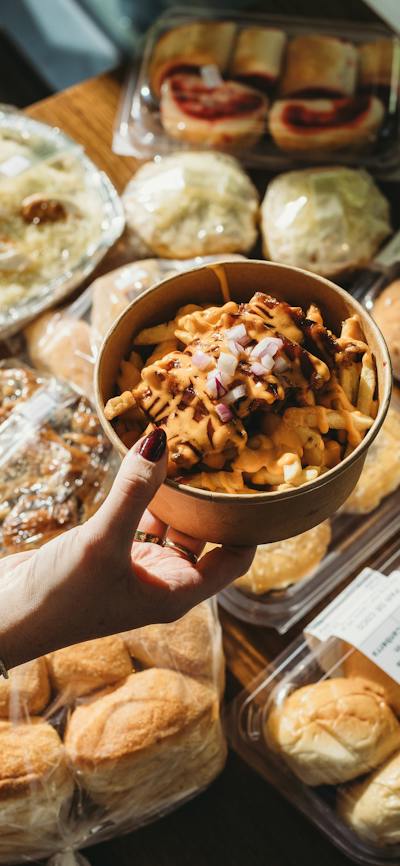 A hand holding a bowl of lechon loaded fries topped with cheese and onions, surrounded by assorted F