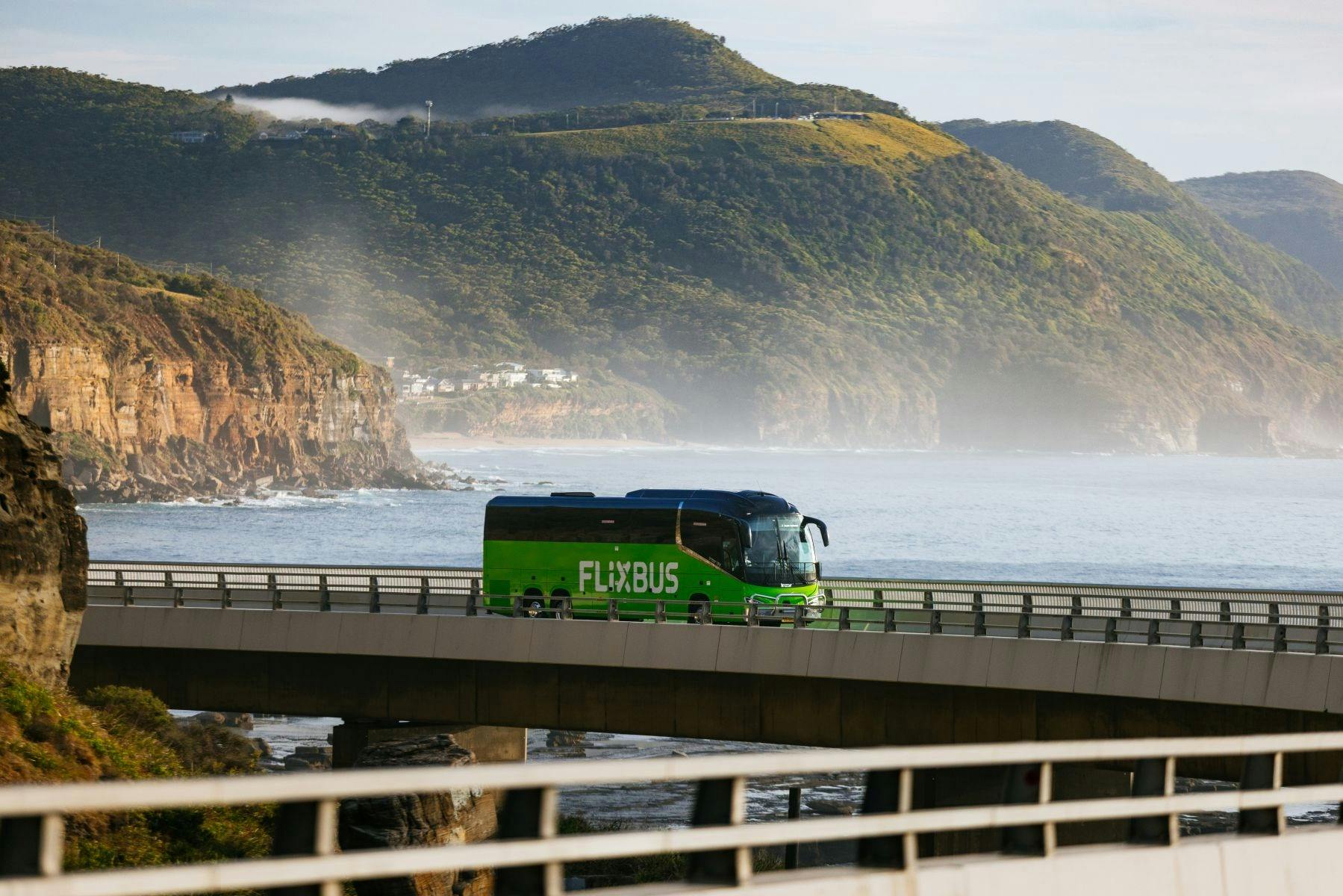 A green flixbus driving on a bridge with sea and mountains in the background.