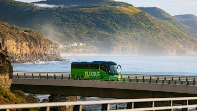 A green flixbus driving on a bridge with sea and mountains in the background.