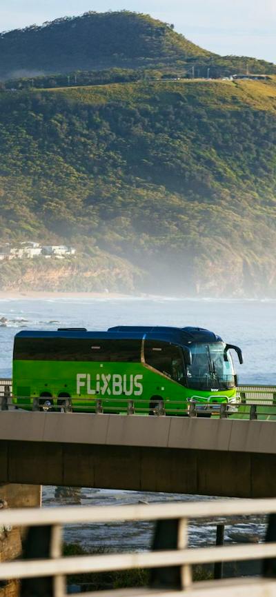 A green flixbus driving on a bridge with sea and mountains in the background.