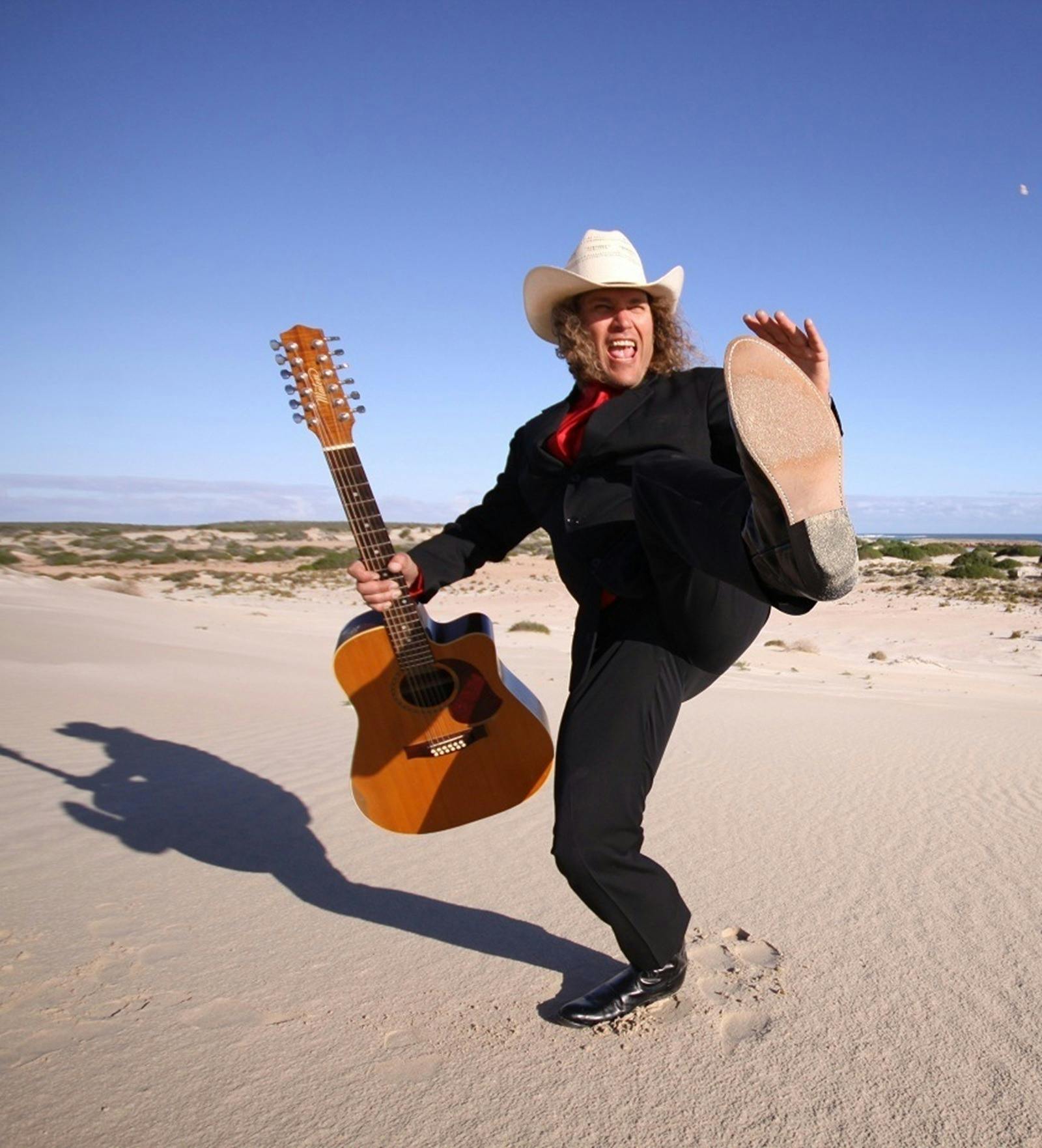 A guy wearing a suit and hat, holding a guitar and kicking his foot out on the beach