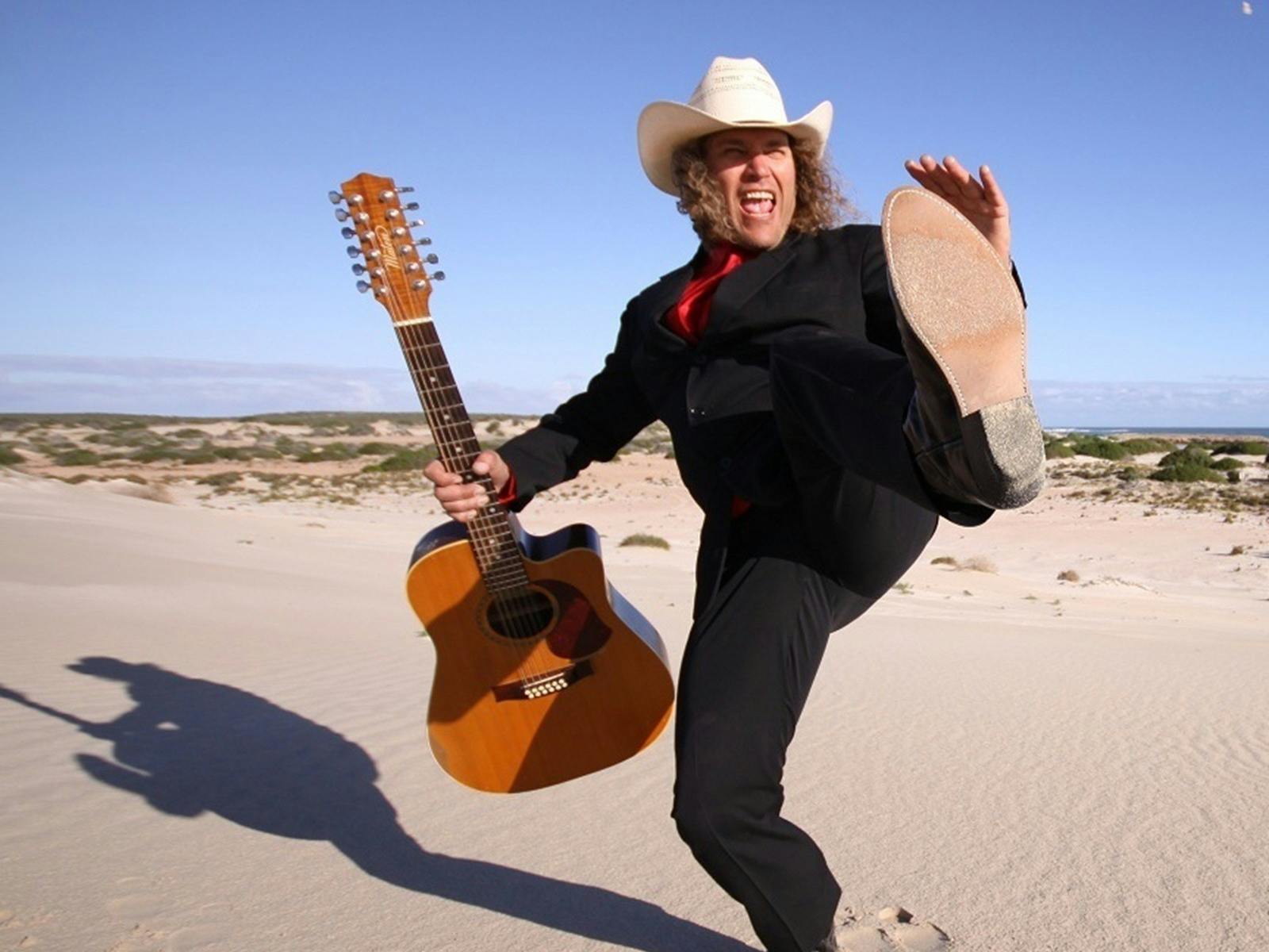 A guy wearing a suit and hat, holding a guitar and kicking his foot out on the beach