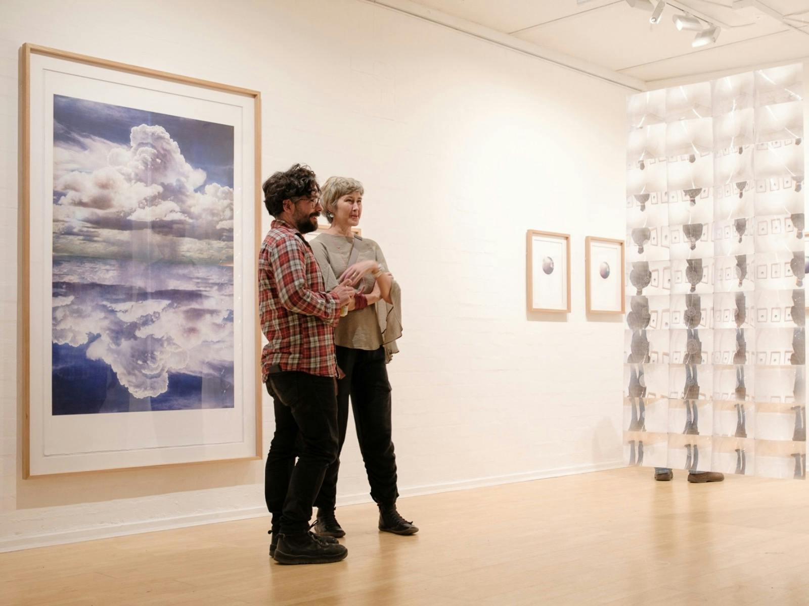 Two people viewing artwork in a bright contemporary gallery, with framed prints and an installation.