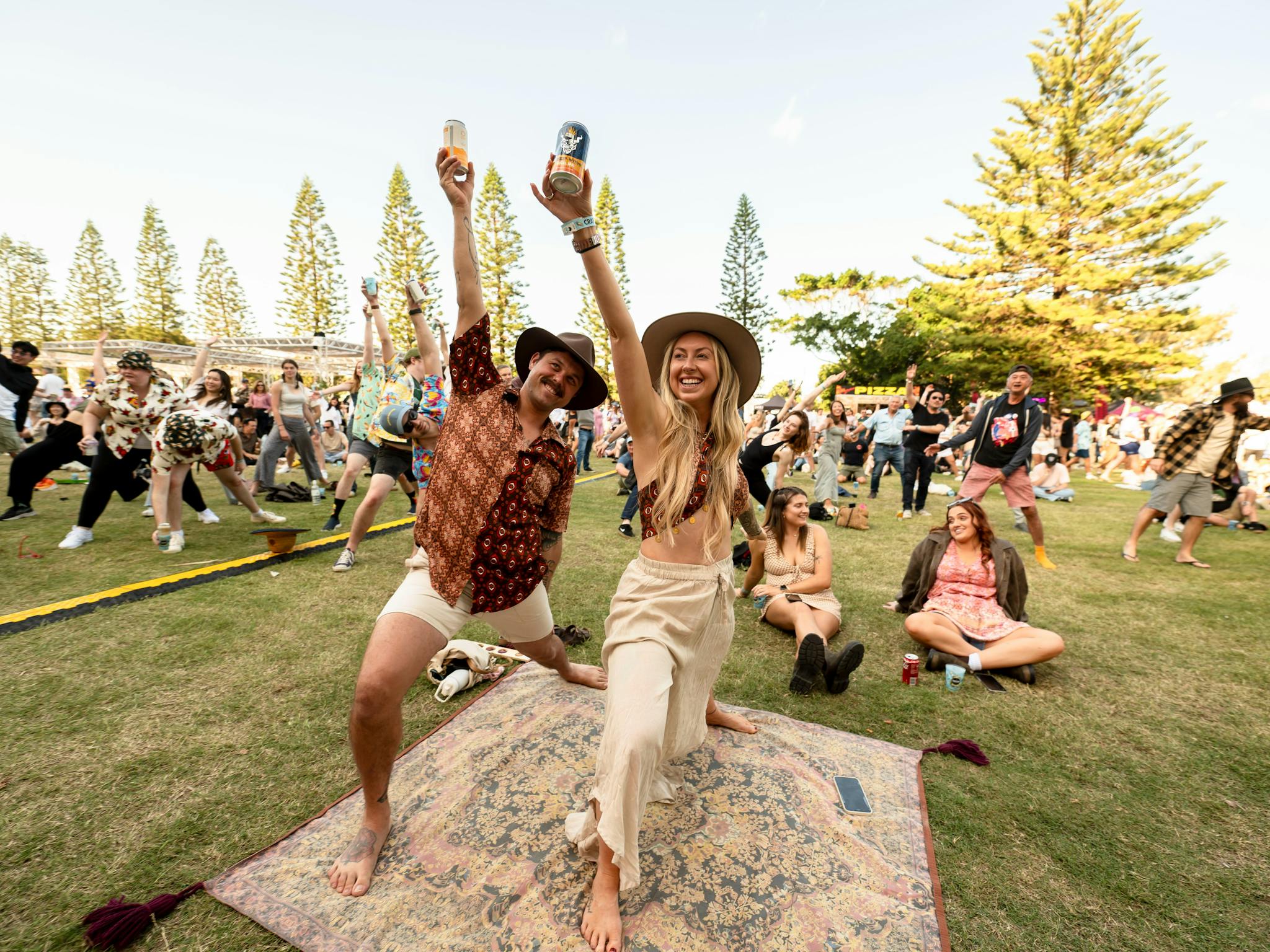A couple stand on a picnic rug holding beers in the sky in a yoga pose
