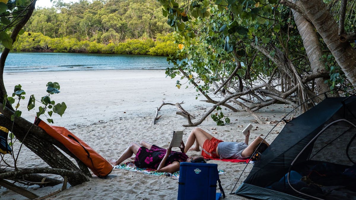 Two people read books on the beach in front of their tent under the shade of tropical trees