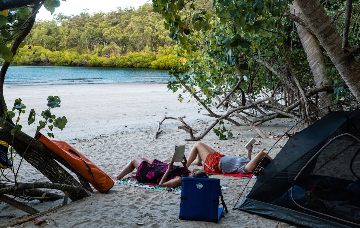 Two people read books on the beach in front of their tent under the shade of tropical trees