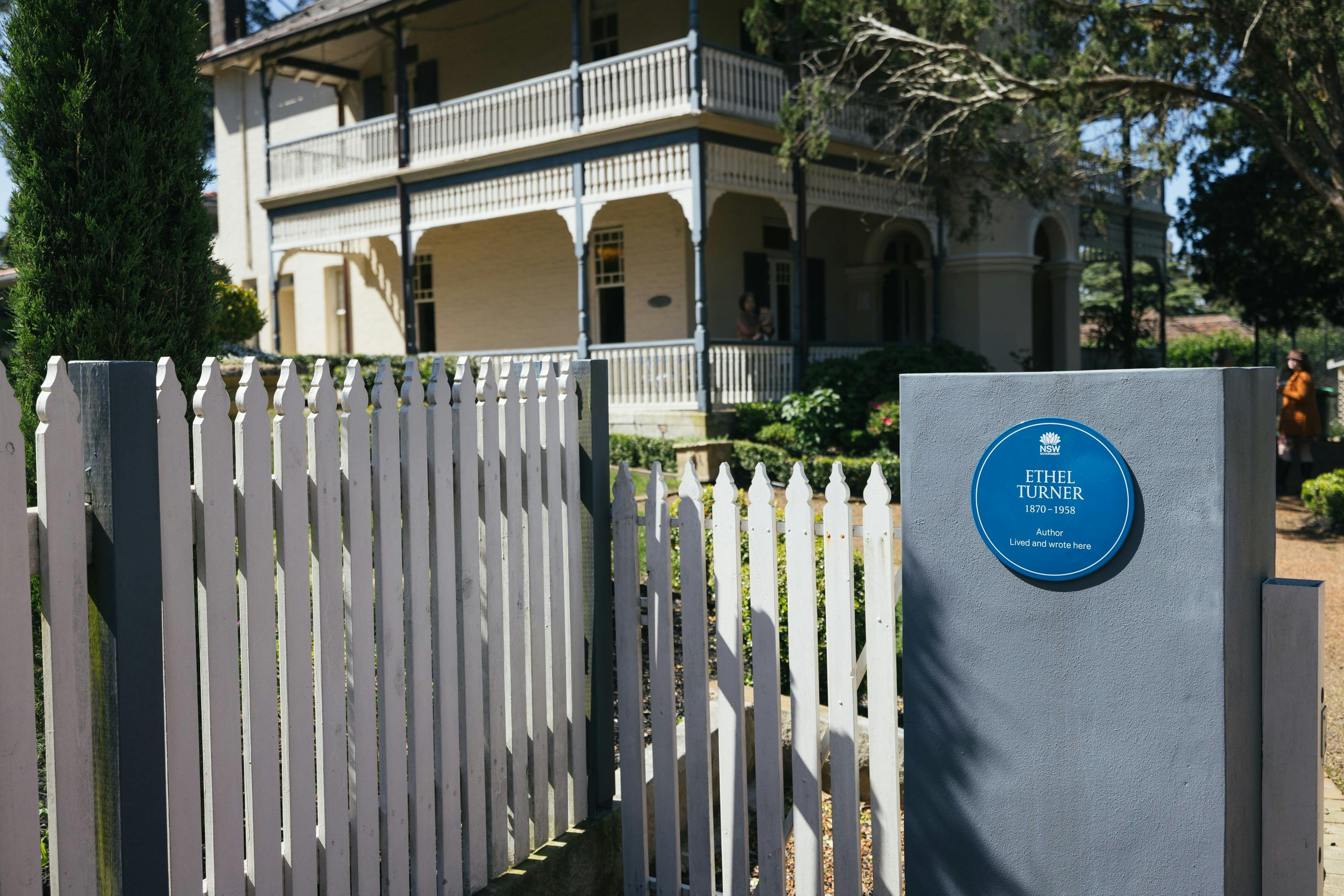 Blue plaque on pillar, white picket fence, two-story Federation Filigree style house in background